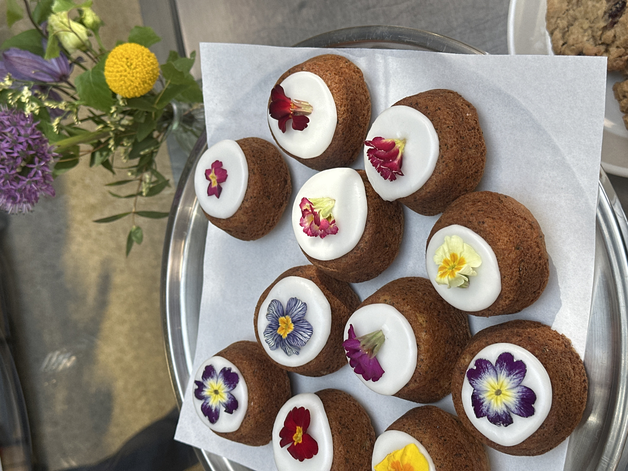 An overhead, close-up shot showcases a collection of artfully decorated mini cakes, likely friands or small muffins, arranged on a silver tray lined with white parchment paper. Each round, dark brown cake is topped with a semicircular dollop of pristine white icing, adorned with a delicate, vibrant edible flower. The edible flowers display a variety of colors, including purple and yellow pansy-like blossoms, deep red, fuchsia, and pale yellow petals, adding a fresh and colorful touch to each treat.
To the left of the tray, a vibrant floral arrangement adds to the aesthetic. This bouquet features striking spherical purple allium flowers, elegant purple clematis blooms, and a bright yellow spherical craspedia (billy button) flower, all interspersed with lush green foliage. The background is softly blurred, hinting at an outdoor or well-lit indoor setting, possibly a cafe or market stall in London, United Kingdom, given the provided context.
The scene conveys a sense of delicate artistry and fresh ingredients, possibly for sale or display at a bakery, event, or special occasion. The bright, natural lighting suggests it is daytime. There is no visible text in the image.