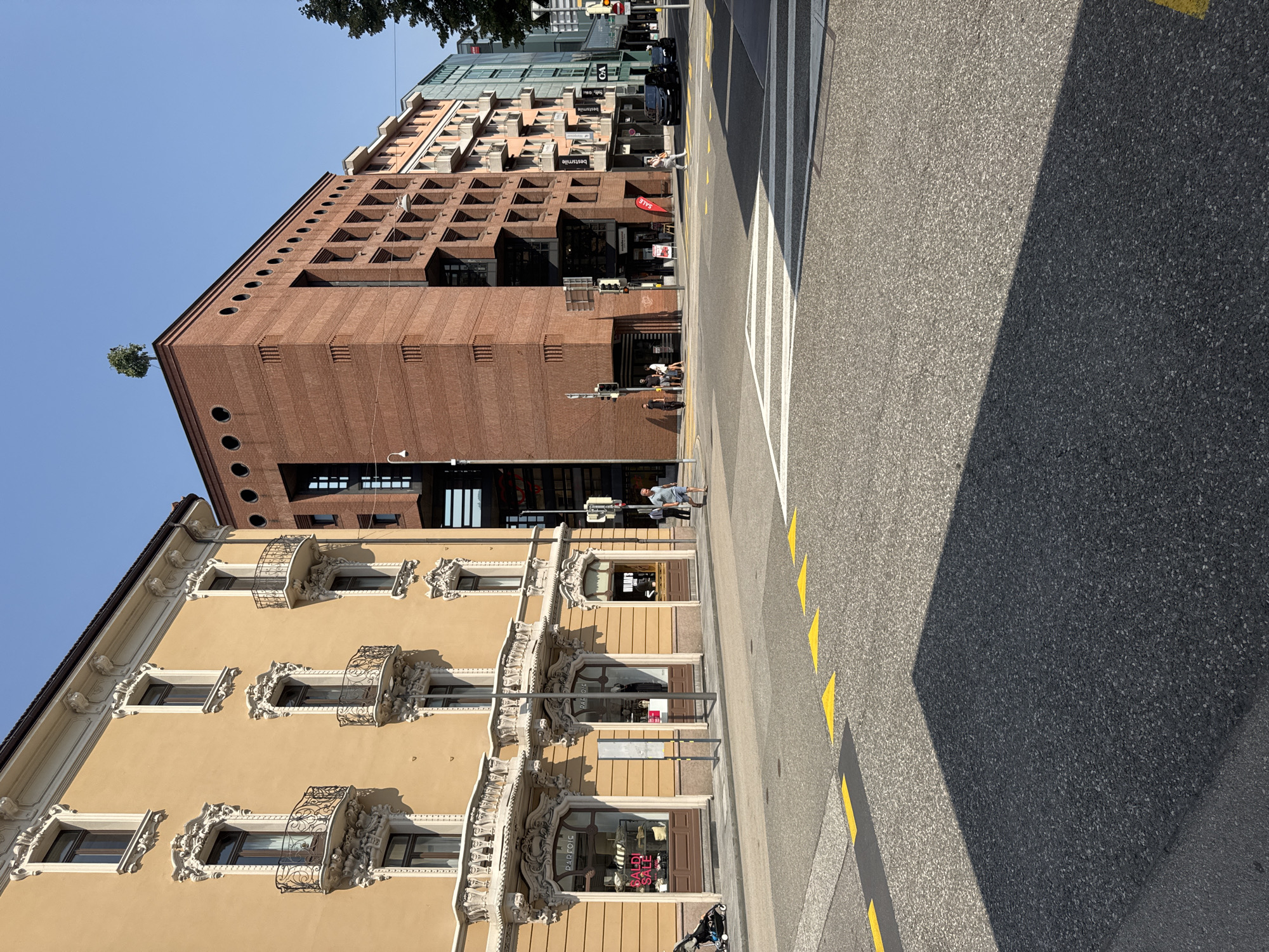 The image shows a street scene in Lugano, Switzerland. In the foreground, a crosswalk with yellow painted triangles is visible on the asphalt road, with a large shadow cast across it. To the left, a building with a light yellow facade and ornate white balconies is prominent. Its ground floor features shopfronts with signs for "PARFOIS" and "BALDI SALE." A stroller is parked on the sidewalk in front of the building.
To the right of the yellow building, a modern, red brick building with circular windows and a distinctive facade rises. A small tree is planted on its rooftop. Further to the right, a series of apartment buildings with balconies and a glass-fronted building are visible, with signage for "best smile" and "C&A" on them.
Several people are present in the scene, including a few individuals walking on the sidewalks and a man in a blue shirt and shorts walking across the street. A black car is parked near the modern brick building. The sky is clear and blue, indicating a sunny day. The overall environment suggests a commercial and residential area in a European city.
