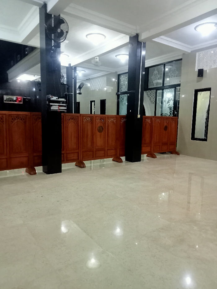 The image shows the interior of a mosque in Kecamatan Margorejo, Indonesia, with a white marble floor. The room is empty with a wooden partition in the foreground, and two black pillars in the background. The mosque is well-lit with multiple ceiling lights and windows. There is a large window with decorative glass and a smaller window with a patterned white film. A ceiling fan can be seen in the top left corner. The room is clean and empty, with no people present.