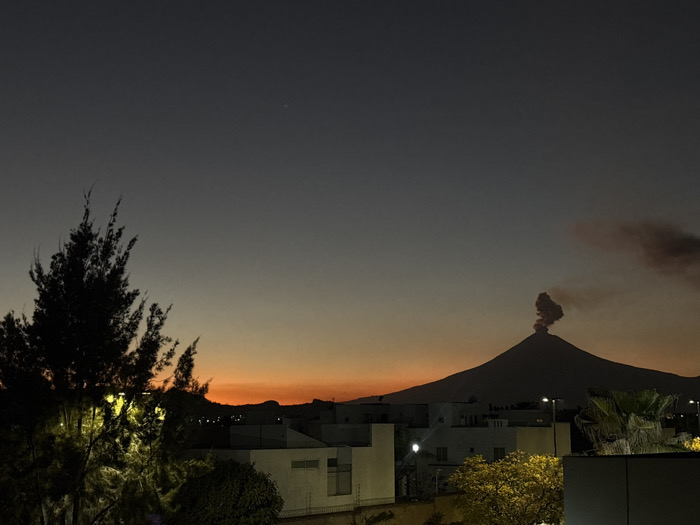 A dusky landscape features a silhouetted volcano emitting a plume of smoke, set against an orange and gray twilight sky. Buildings and trees, some illuminated by streetlights, occupy the foreground. A dark evergreen tree stands to the left. The overall scene suggests a tranquil yet potentially volatile setting.