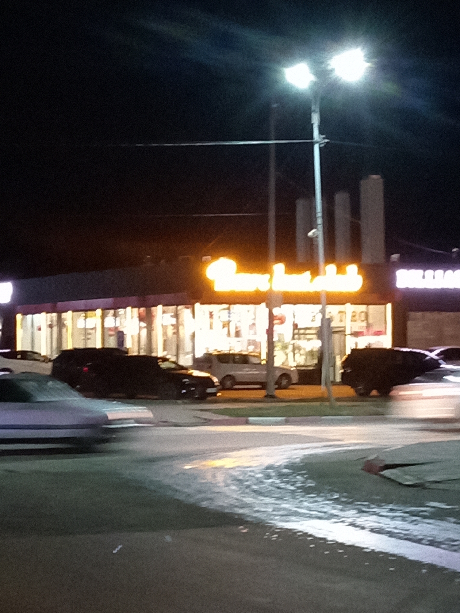 The image depicts a nighttime scene outside a commercial building in Rudny, Kazakhstan. A bright streetlight with two bulbs illuminates the area. The building's facade features large windows and prominent signage. One sign, illuminated in orange, appears to be in Arabic script, spelling out "Burger Khasan". To the right, another sign with white lettering is partially visible. Several vehicles are present: a black SUV parked in front of the building, a light-colored sedan moving in the foreground, and another dark vehicle further down the street. The ground appears wet and reflective, possibly from recent rain or dew. The overall impression is that of a commercial establishment operating at night.