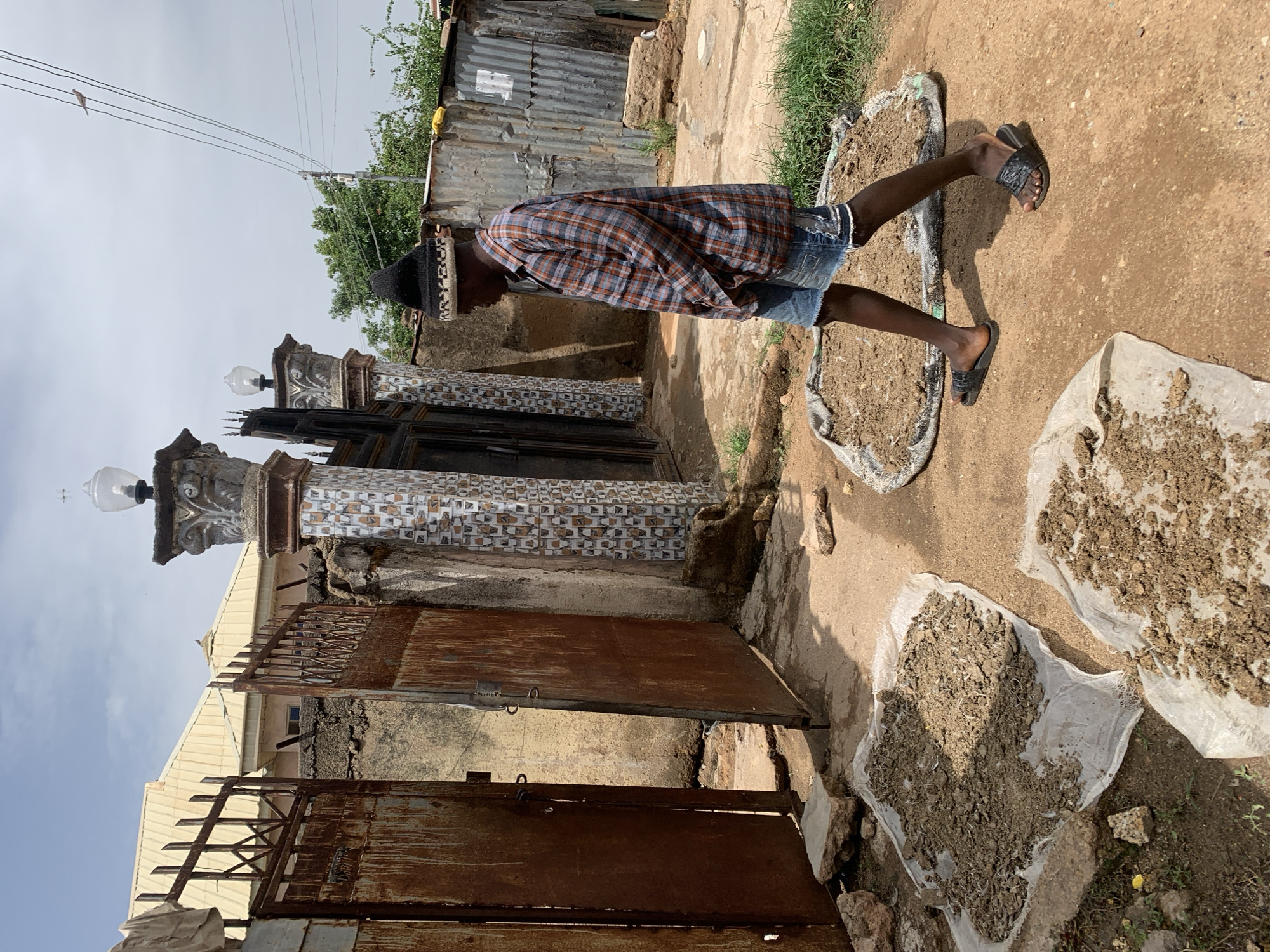 The image depicts a person in casual wear standing near three piles of drying, processed earth in what appears to be a residential yard in Jos, Nigeria. The person, wearing a plaid shirt, denim shorts, and sandals, is also wearing what appears to be a skull cap. The yard is adjacent to a property enclosed by rusty metal gates and decorative pillars with light fixtures. The drying earth is laid out on white plastic sheeting and in what appears to be the torn edges of a bag. In the background, corrugated metal fencing and vegetation are visible. Overhead power lines are also visible in the background. The area appears to be a mix of paved and unpaved surfaces.