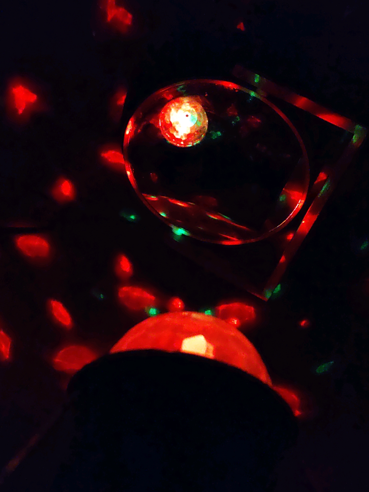 The image shows a close-up, dark-toned photograph with red and green lights reflecting off a dark background. In the foreground, a dark, rounded object, possibly a speaker or a light fixture, is visible, topped with a bright red light. Above and to the right, a clear, rounded glass surface reflects a bright, multicolored light. A thin, angular, red-lit object, possibly a frame or a reflective surface, is positioned behind the glass. The background is filled with numerous diffused red and green spots of light, suggesting a disco ball or a similar lighting effect in a dimly lit environment. There are no people or discernable city elements visible. The location of Rostov-on-Don, Russia is not depicted or suggested in this image.