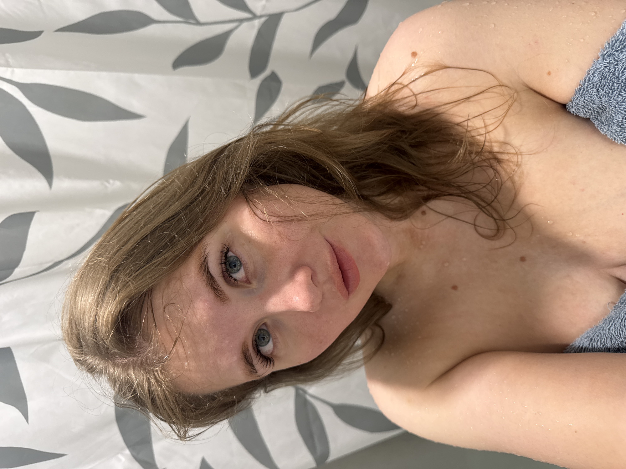 A woman is in a shower, wrapped in a towel, with water droplets visible on her face, hair, and body. The shower has a white and gray leaf-patterned curtain. The location is not discernible as being in Ферма, Russia, based solely on this image.