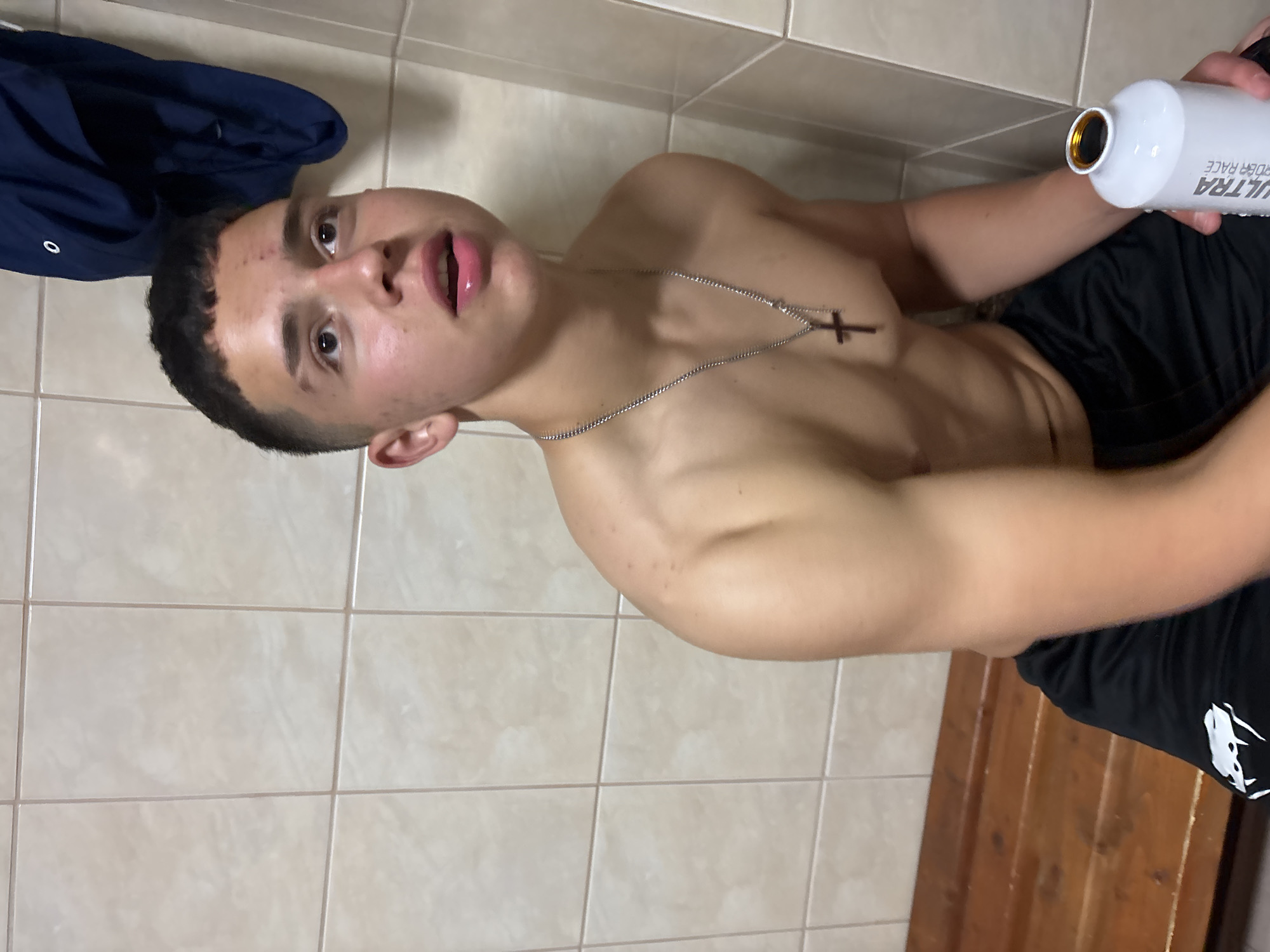 A shirtless young man is seated indoors against a tiled wall. He is holding a white water bottle. A cross necklace hangs on his chest. He is wearing black shorts. A blue jacket is partially visible in the upper left. The scene takes place within a locker room. No specific landmarks indicating Zlatograd, Bulgaria, are present.