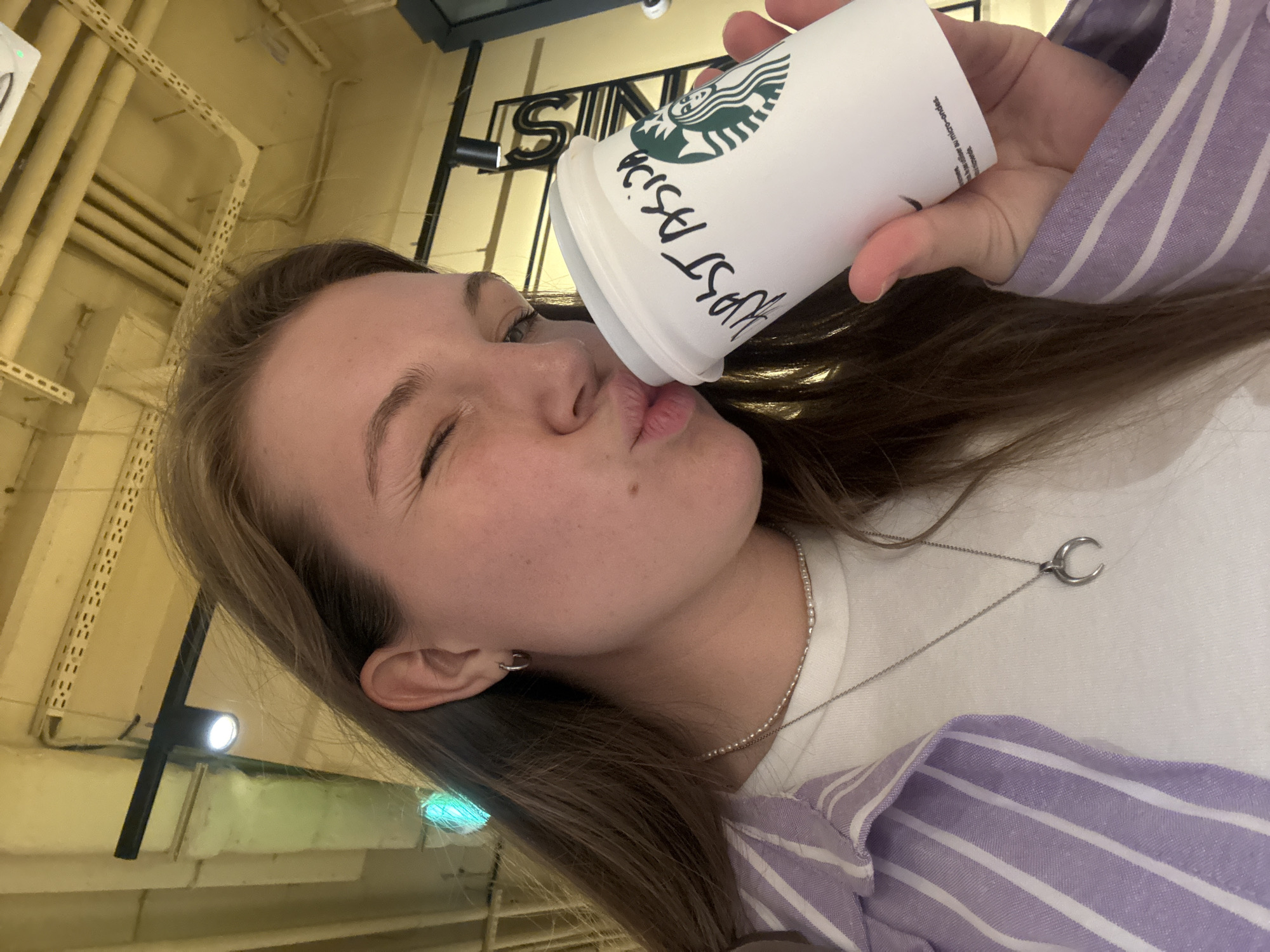 A young woman with light skin, brown hair, and freckles puckers her lips towards a white Starbucks coffee cup. The cup has the Starbucks logo and writing in black marker. She winks her left eye and wears a white t-shirt with a silver necklace and a purple and white striped shirt over it. The background shows interior architectural details with yellow piping, suggesting the location might be a modern interior space. The exact location within Vracar Urban Municipality, Serbia, cannot be determined from the image.