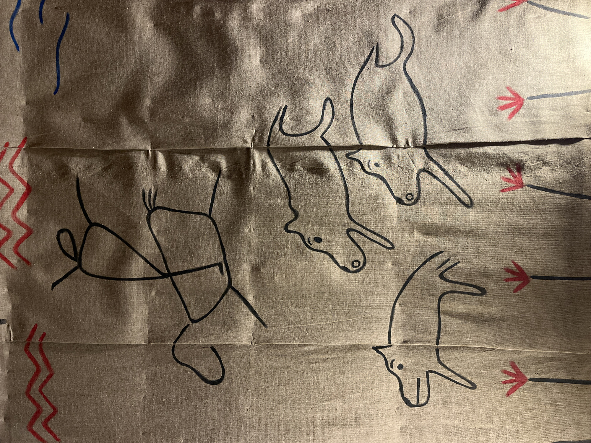This image, located in Saint Petersburg, Russia, displays a stylized artwork painted on a light brown, coarsely woven fabric exhibiting vertical creases. The artwork features black-outlined figures and shapes, accented with red and blue elements. Centrally positioned in the upper half, a stylized human figure is depicted astride a quadrupedal animal, likely a horse, both rendered in outline. Below this, three additional quadrupedal animals, each with simple dot eyes and a snout, are shown in active running or leaping poses. The upper left includes a horizontal band of red zigzag lines, while the upper right shows two horizontal blue wavy lines. Along the bottom, several stylized red flowers with black stems are arranged. The scene is evenly lit, accentuating the fabric's texture and the drawn elements.