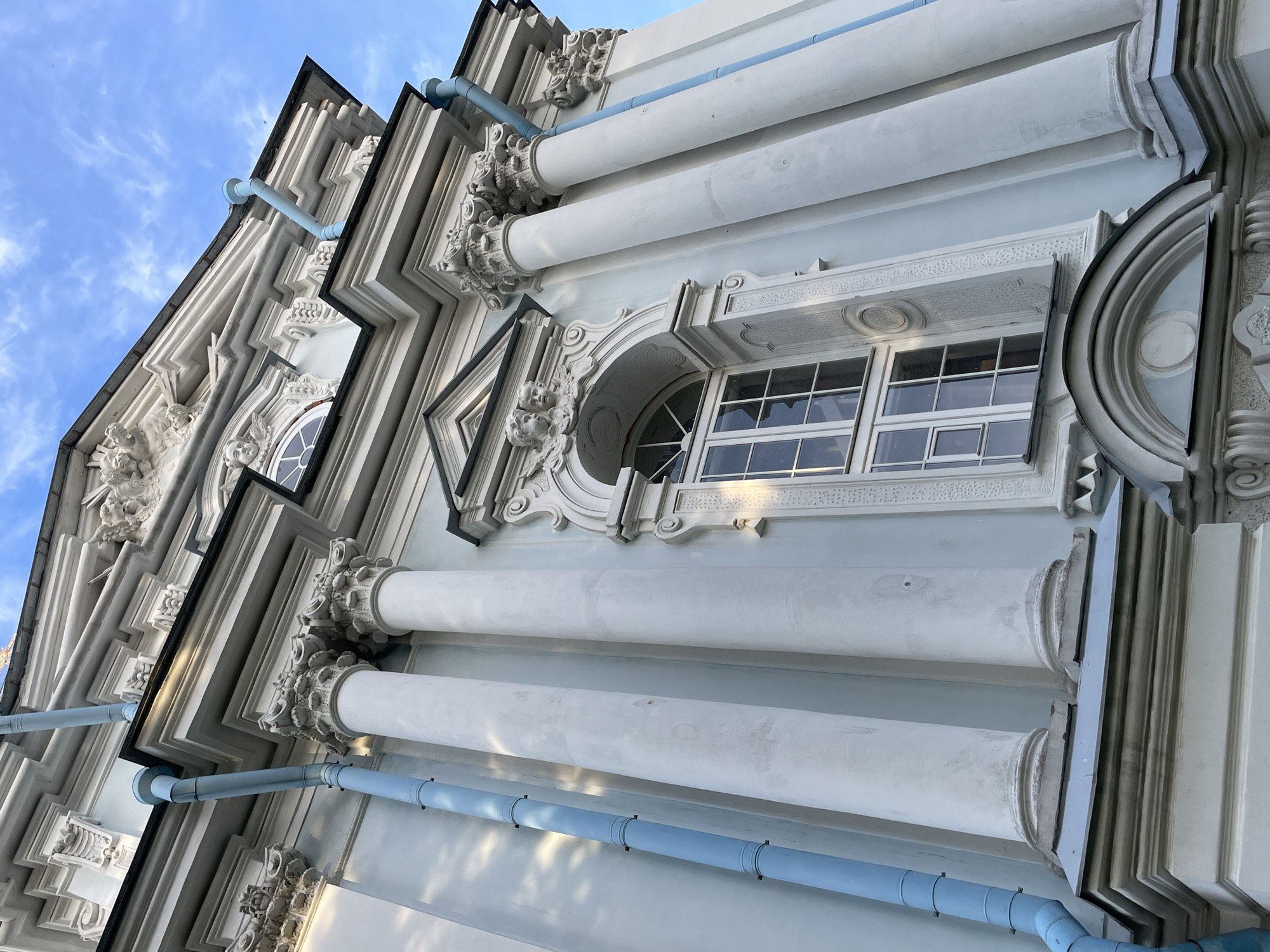 The image presents a low-angle, upward-looking view of an ornate architectural facade, situated in Unknown, Russia. The building's exterior is characterized by a light blue-grey primary color, contrasted with extensive white architectural details. Multiple tall, cylindrical columns with elaborate, possibly Corinthian-style, capitals are prominent, supporting a series of white cornices and friezes. One large, arched window with a white frame and multiple glass panes is centrally visible, flanked by columns. Above this window, intricate carvings, including two sculpted figures resembling putti or cherubs, are integrated into the decorative stonework. Additional decorative moldings, architraves, and a smaller, semicircular archway are present at the base of the visible structure. Two light-blue vertical drainpipes are affixed to the facade, one on the far left of the frame and another visible on the right, extending downwards from the roofline. The upper sections of the building and roofline are silhouetted against a clear blue sky with scattered white clouds. Sunlight illuminates the building from the right, casting soft shadows on the left portion of the facade. No individuals or active subjects are present in the scene.