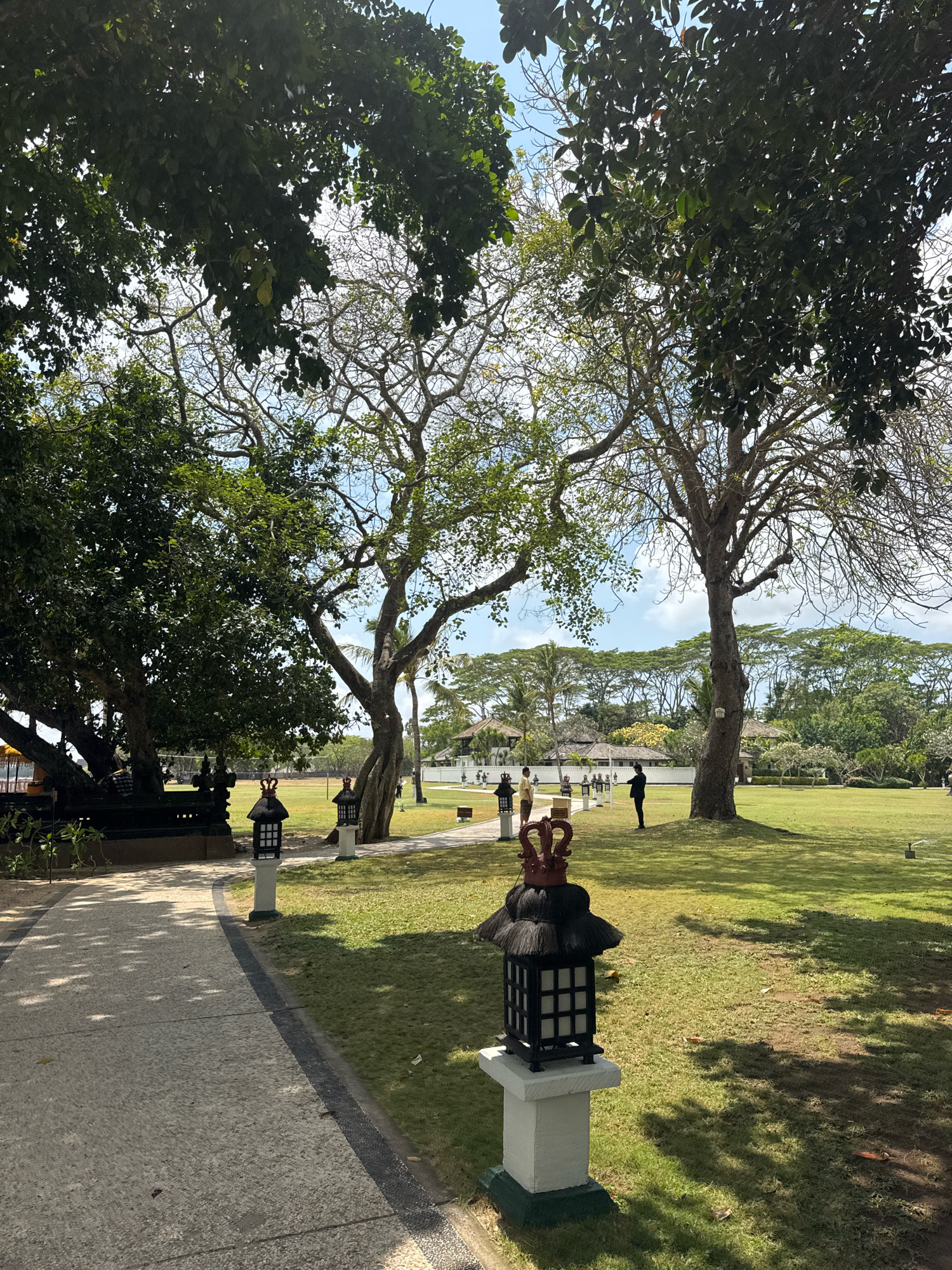 This image depicts a sunlit, outdoor scene within what appears to be a temple complex or resort in Kuta Selatan, Indonesia. A paved walkway, lined with decorative lanterns and lush green grass, curves through the frame. In the background, several individuals are visible, some standing and observing, while others are engaged in conversation. Large, mature trees with expansive branches dominate the foreground and midground, casting dappled shadows across the landscape. The architecture in the distance suggests traditional Indonesian design, with tiled roofs and white walls. The sky is a clear blue, with scattered clouds, indicating pleasant weather. The overall atmosphere is serene and tranquil.