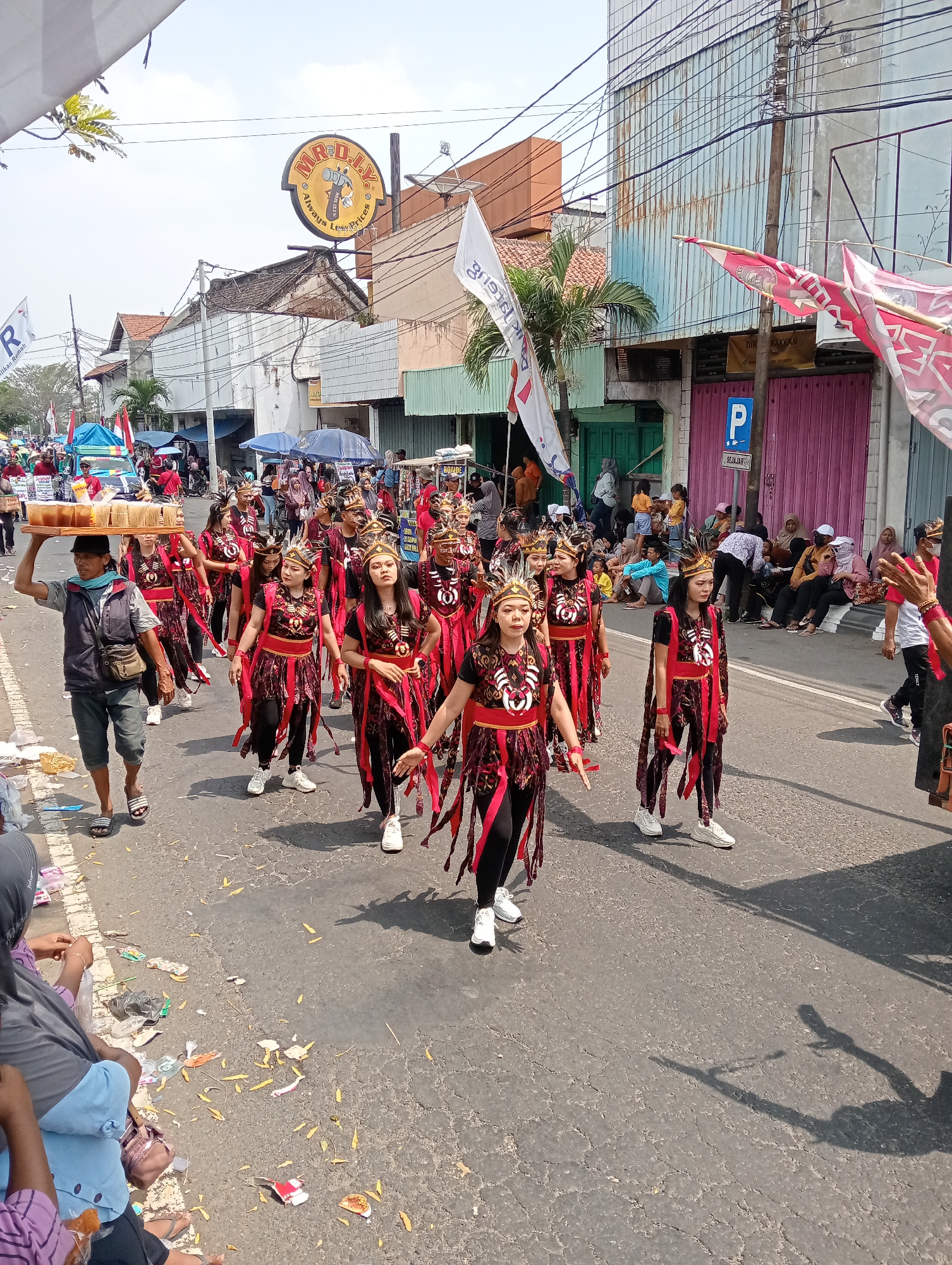 A group of dancers in traditional attire with black and red costumes, ornate headwear, and intricate jewelry are parading down a paved street in Demak, Indonesia. The dancers are performing in unison, with synchronized arm movements. A man is walking ahead of them, carrying a tray of baked goods. Spectators are gathered on both sides of the street, some sitting on the curb and others standing. Flags and banners are visible, along with various shops and buildings lining the street. The weather appears to be sunny.