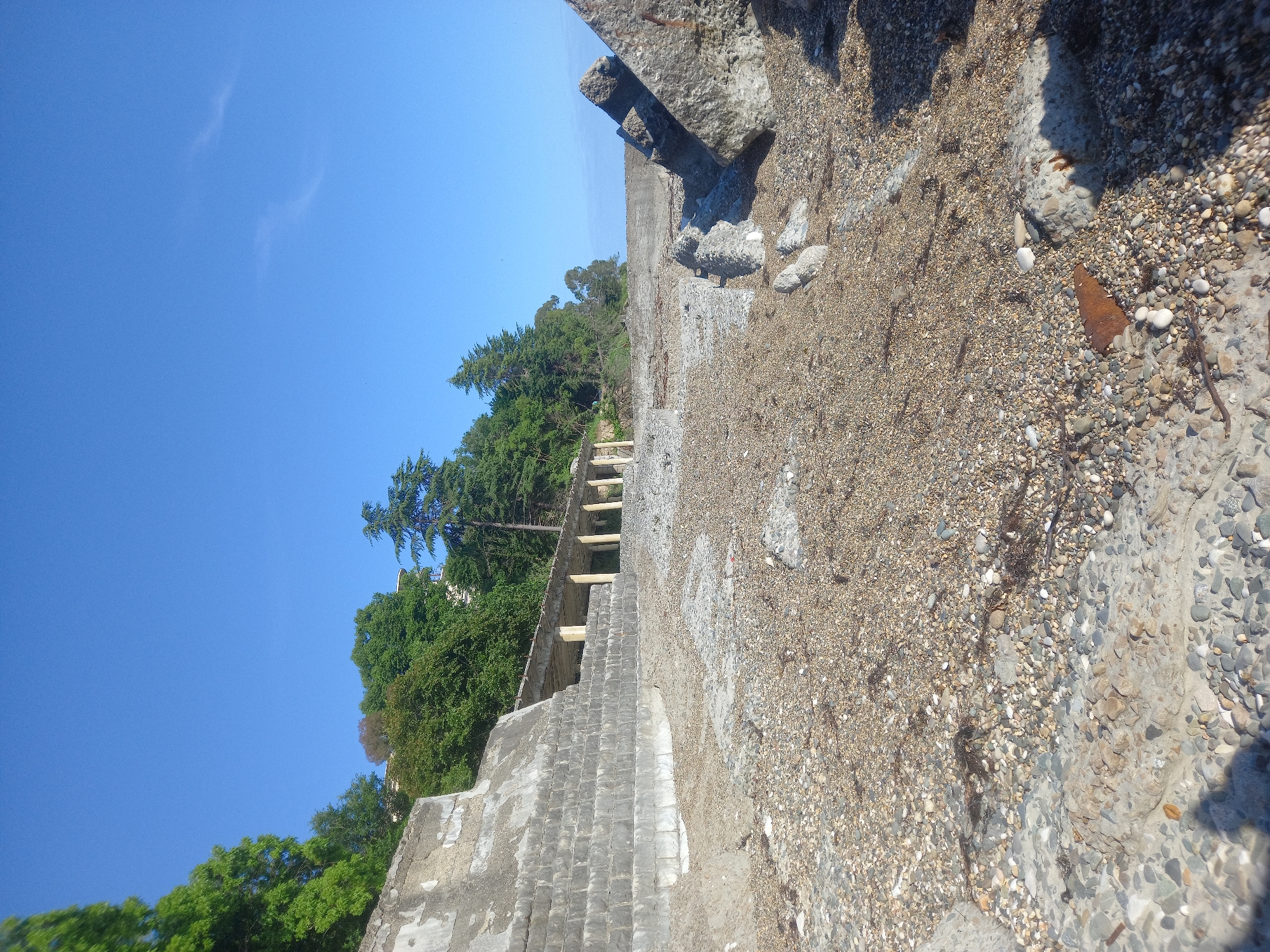 The image depicts a rocky shore with a clear blue sky and some scattered white clouds. The foreground is dominated by a grey, weathered concrete structure with a textured surface of small stones. To the left of the concrete structure, there is a lush green hillside covered with trees. A partially visible set of wooden stairs or a ramp leads up the hillside. The overall scene suggests an outdoor setting, possibly a coastline or a body of water. The time of day appears to be daytime, given the bright sunlight and clear sky. There is no visible text in the image. The orientation of the image is tilted.