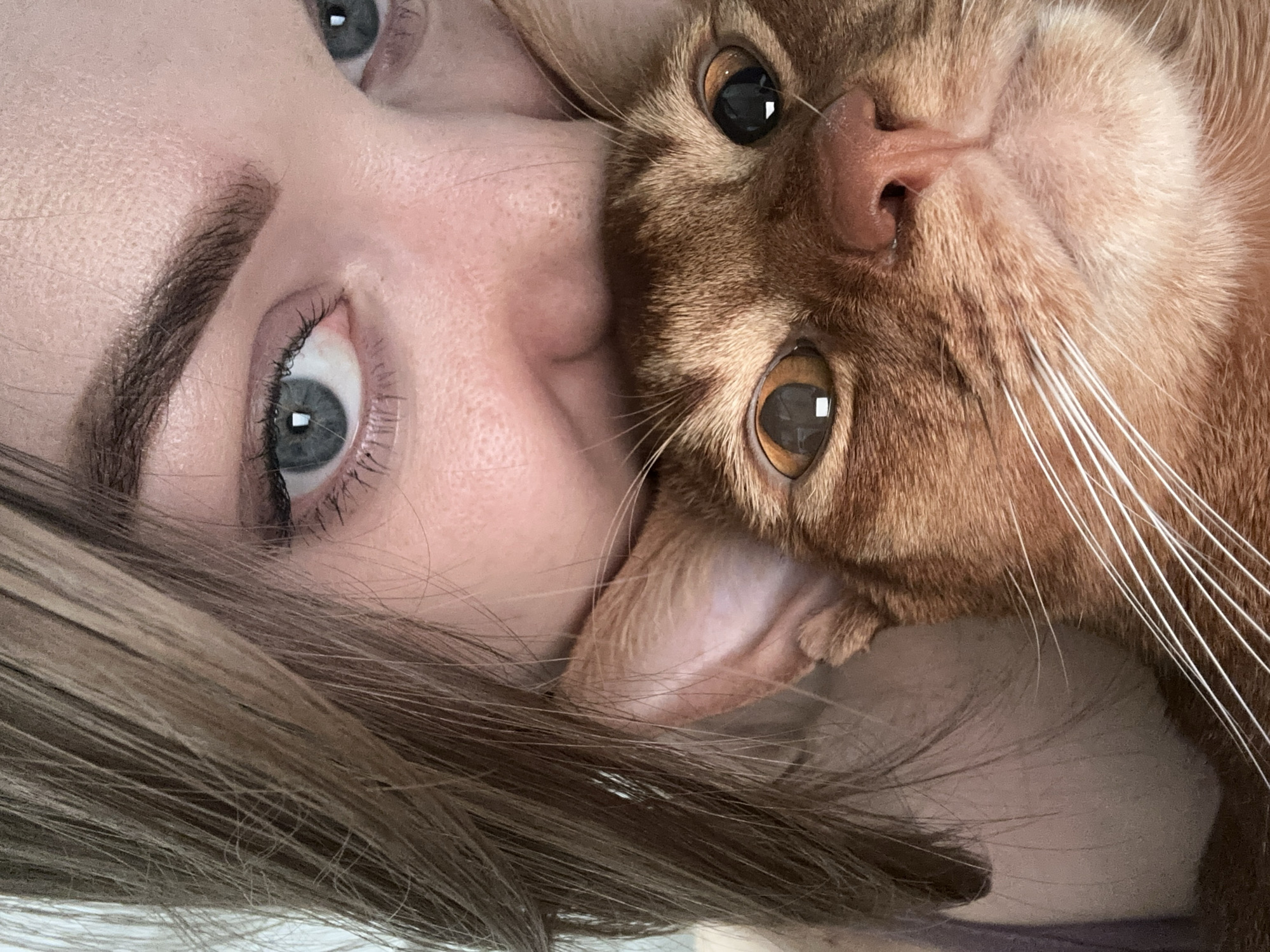 A close-up, selfie-style image features a woman and an orange tabby cat. The woman's face is tilted upwards, with her blue-gray eyes and a portion of her eyebrow visible. Her light brown hair is draped around her face. The cat is positioned closely to the woman, its face appearing upside down. The cat has amber eyes, a pink nose, and long white whiskers. The cat's ear is visible near the woman's cheek. The background is out of focus. There is no indication the image was taken in Kazan, Russia.