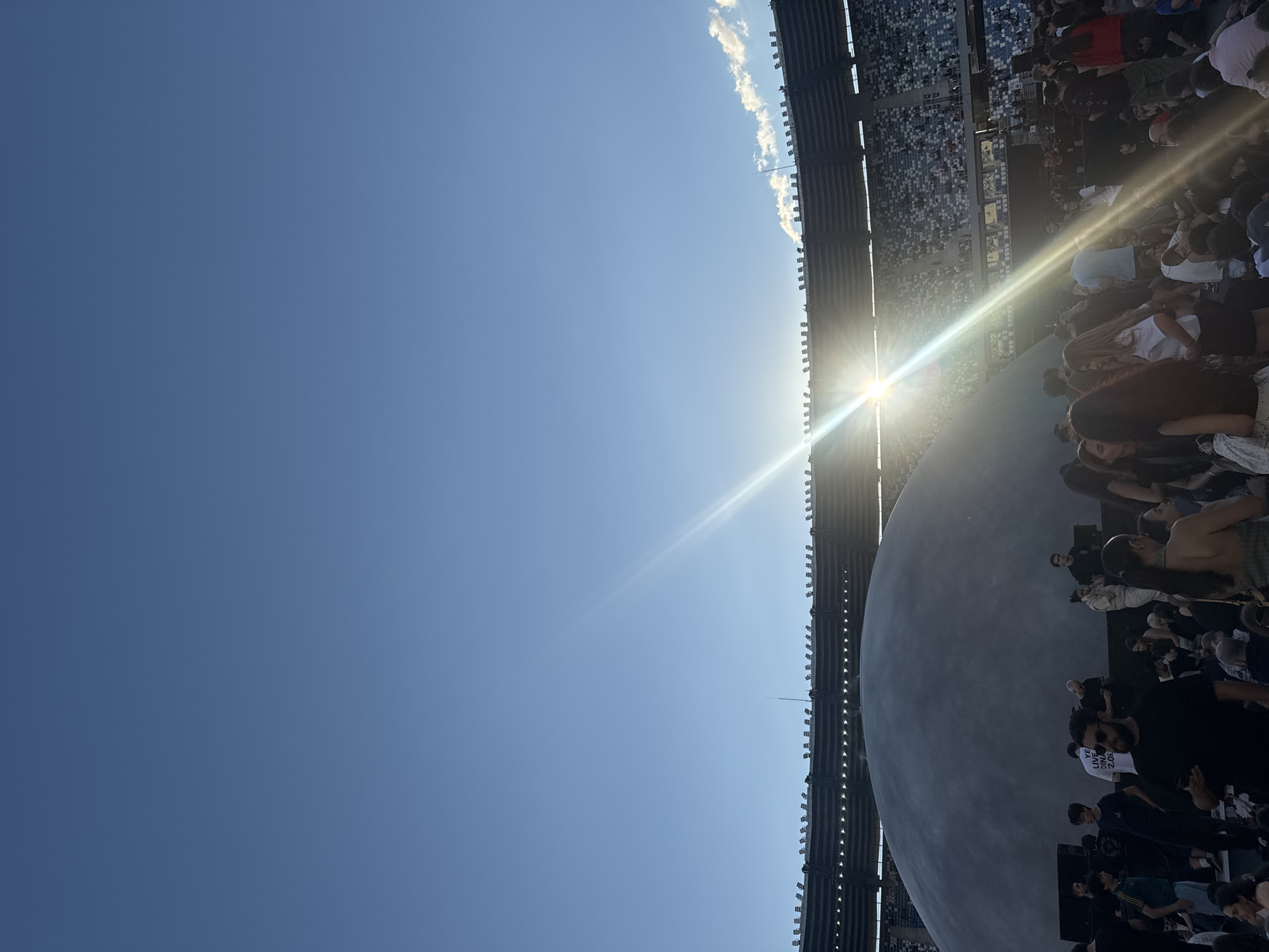 The image depicts a large gathering of people outdoors, with a stadium structure in the background. The sky is clear and blue, with the sun shining brightly, creating lens flare across the scene. In the foreground, a large, grey, dome-like structure is visible. A crowd of individuals of various ages and genders are congregated around the dome and in the stadium seating. Some people are standing, while others appear to be sitting or observing the surroundings. The overall atmosphere suggests an event or gathering taking place at a venue resembling a stadium in Tbilisi, Georgia. The lighting indicates it is daytime, likely late afternoon or early evening due to the low angle of the sun.