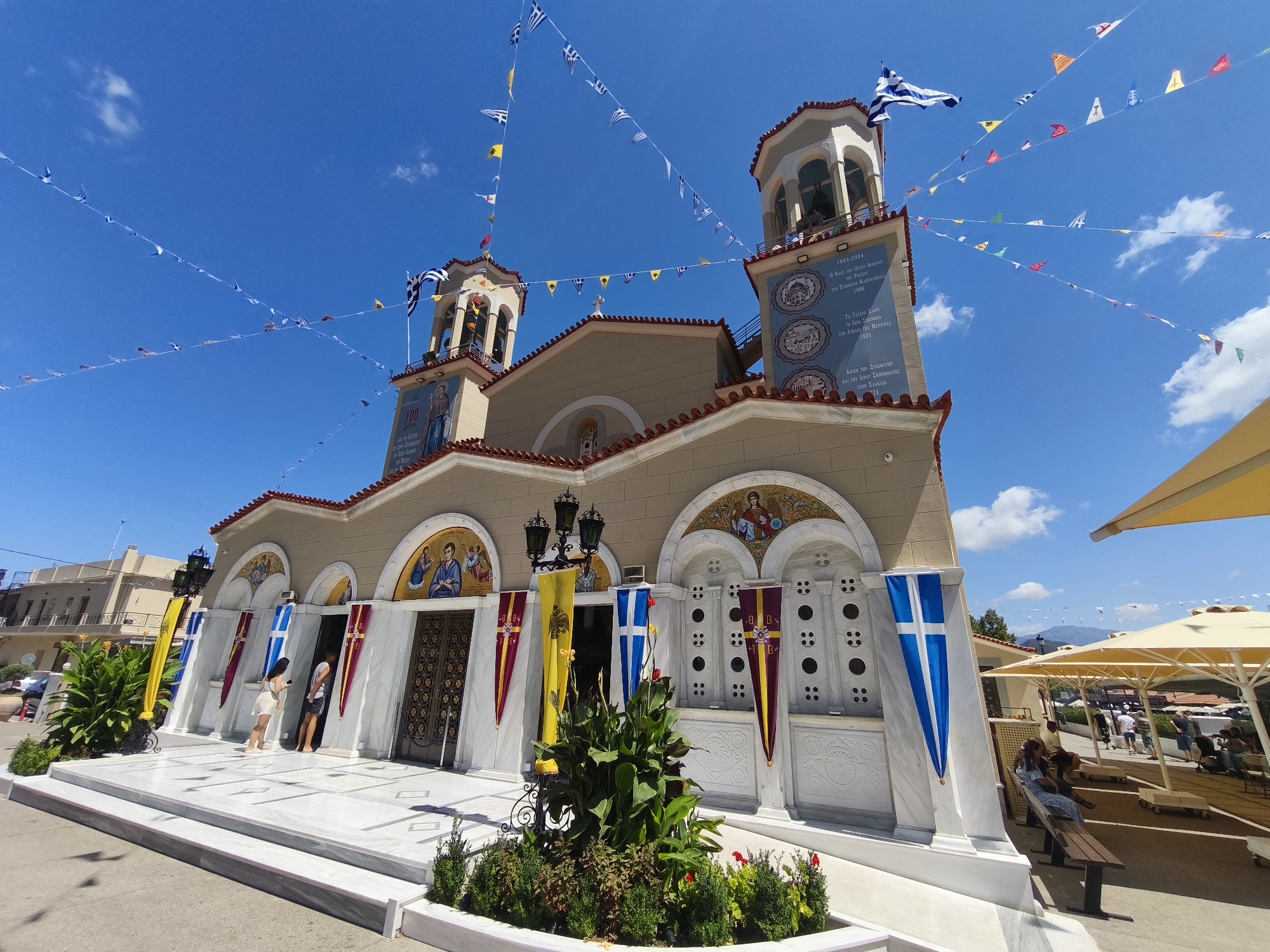A wide-angle, eye-level shot captures the exterior of a Greek Orthodox church in the city of Prokopi, Greece, under a clear blue sky with scattered white clouds. The church, painted a light beige with red-tiled roofs, features two prominent bell towers adorned with Greek flags and illuminated by the bright sun. Bunting flags in various colors, including Greek flags, are strung across the sky above the church.
The church entrance is grand, with double doors and arched mosaics above. The facade is decorated with marble elements, and religious banners hang on either side of the entrance. A lush green bush sits in front of the entrance, adding a touch of nature to the scene. Two individuals, a woman in a white dress and a man in a grey shirt, are standing near the entrance.
To the right of the church, several people are seated under large, cream-colored umbrellas, suggesting an outdoor gathering or seating area. The background reveals more buildings and distant hills, indicating a town or village setting. The overall atmosphere is festive and serene, likely during a religious celebration.