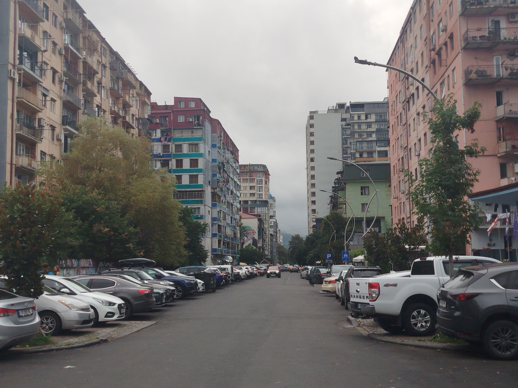 The image depicts a street scene in Batumi, Georgia, characterized by a mix of multi-story apartment buildings and parked vehicles. The buildings on either side of the street are diverse in their architecture and color, with some featuring balconies and air conditioning units. Trees and foliage are interspersed along the sidewalk and between buildings.
Numerous cars are parked along the street, both on the right and left sides, filling most of the available space. The vehicles include a variety of models, such as sedans, SUVs, and pickup trucks. Some cars have roof racks.
In the distance, the street appears to extend towards a more urban or commercial area, with more buildings visible. The sky is overcast, suggesting a cloudy day. There are streetlights visible along the road. A few people can be seen in the background, near the parked cars on the left side of the street. The overall impression is of a residential and urban street in Batumi, likely on a day with subdued lighting.