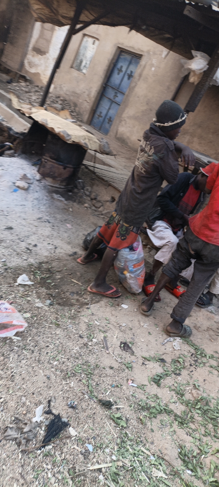Amidst a dilapidated landscape with damaged buildings, a group of children gathers. One wears a beanie, a dark jacket, and red patterned shorts, holding a plastic bag filled with various items. Another child sports a red shirt and dark pants, while a third is partly visible. The ground is littered with debris and sparse vegetation, adding to the overall impression of poverty and neglect.