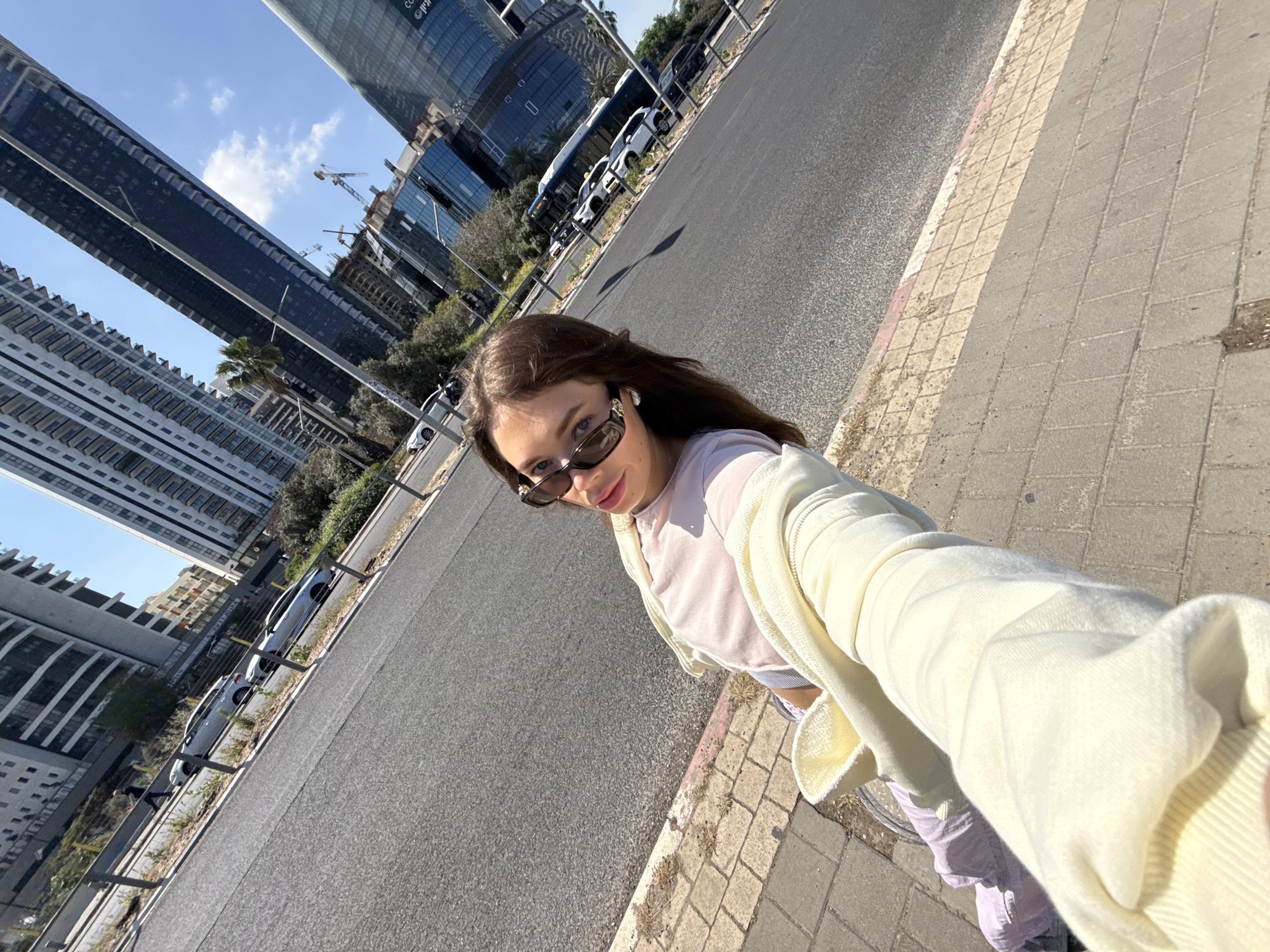 The image is a selfie taken by a woman on a sidewalk next to a street. She is wearing sunglasses, a light pink top, and a pale yellow sweater. The background shows a cityscape with modern high-rise buildings, including some under construction with cranes visible. Cars and buses are parked along the street. The location is likely Tel Aviv, Israel, given the style of architecture. The weather appears sunny and clear.