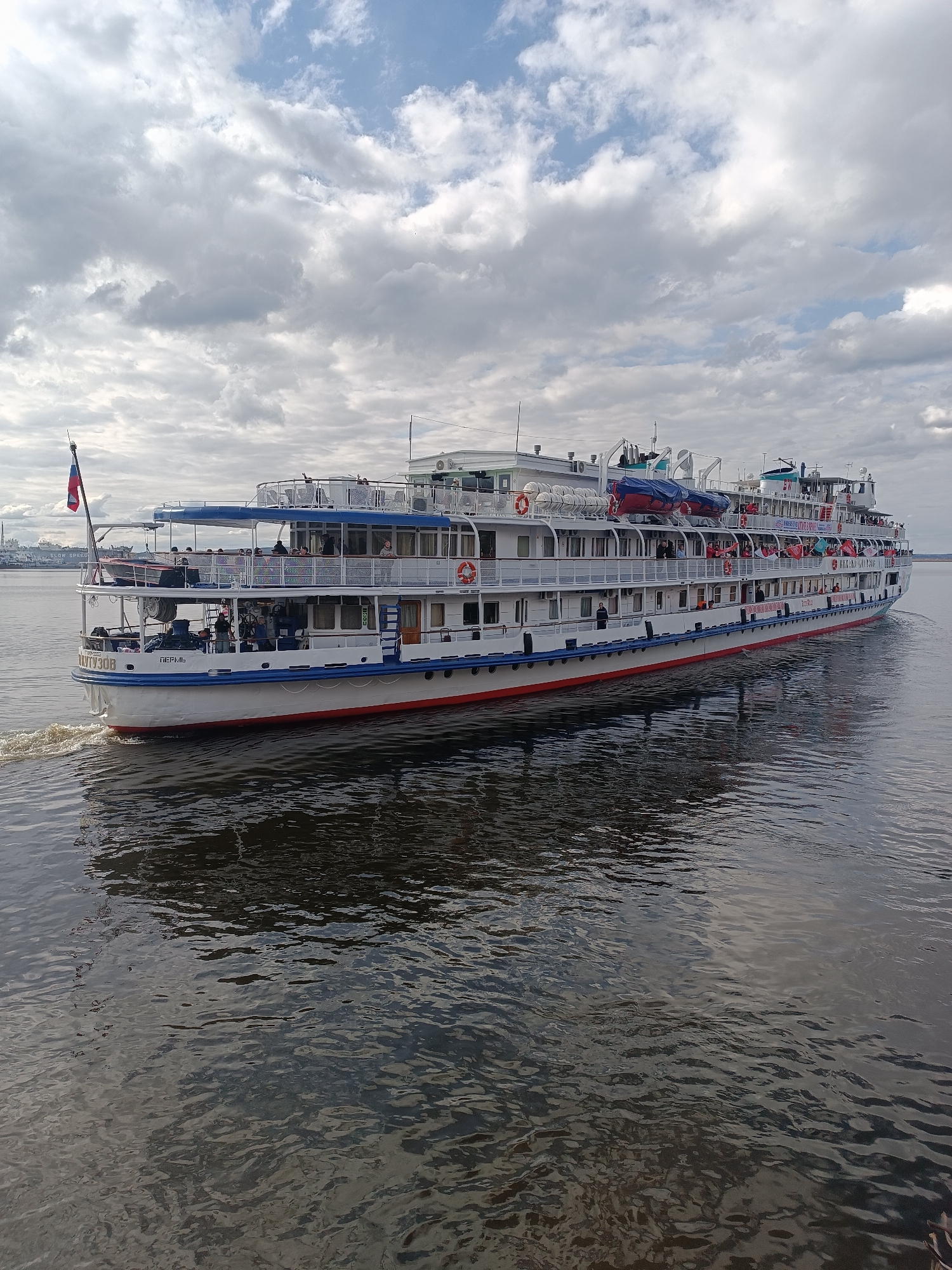 A large, multi-deck river cruise ship, identified as "МИХАИЛ КУТУЗОВ" (MIKHAIL KUTUZOV) with its homeport listed as "ПЕРМЬ" (PERM), navigates a wide body of water. The ship is predominantly white with prominent blue and red stripes along its hull and blue railings on its upper decks. A Russian flag flies proudly from the stern.
Numerous passengers are visible on the open decks of the vessel, standing and observing the surroundings, suggesting a leisure or sightseeing activity. Lifeboats and life rings are stowed along the ship's sides. The ship is actively moving, indicated by the visible wake trailing behind its stern.
The scene is set under a partly cloudy sky, with large, fluffy grey clouds dominating but also showing patches of brighter blue sky. The lighting suggests it is daytime, possibly late morning or afternoon. The water is dark and calm, reflecting the ship and the sky with gentle ripples. In the distant background to the left, faint outlines of land or industrial structures are visible on the horizon. The overall atmosphere appears tranquil and leisurely, typical of a river cruise experience in an area like Chaikovsky, Russia.