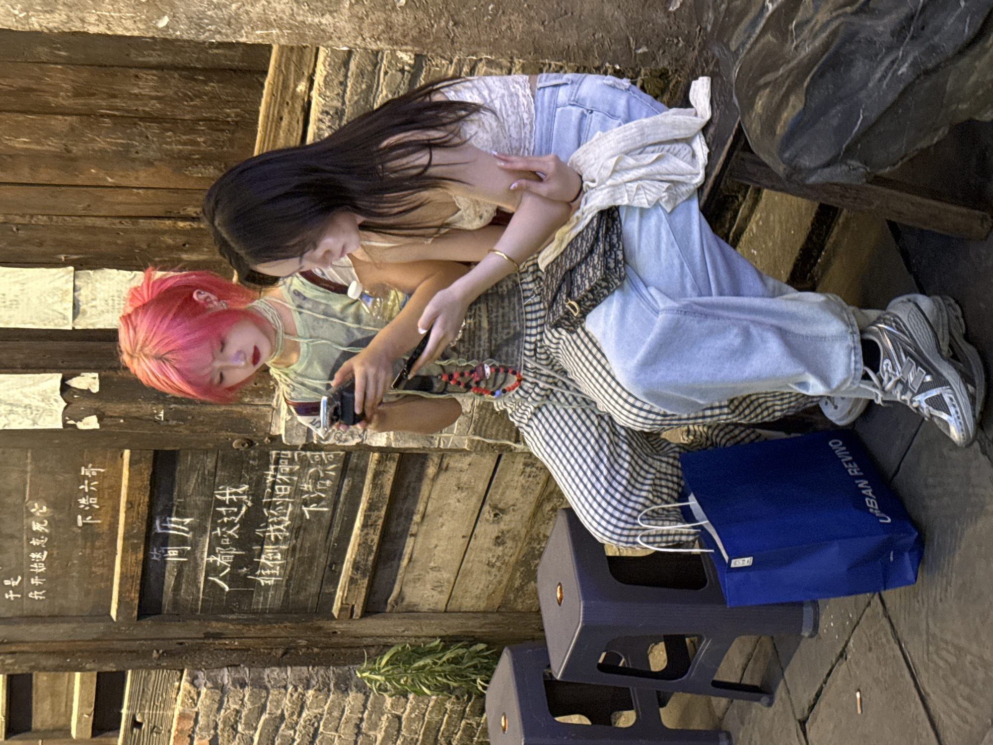 Two young women are sitting outside a building in Nan'an District, Chongqing, China. One woman has long black hair and is wearing jeans and a white top. The other woman has pink hair and is wearing a green top and checkered pants. They are both looking at a phone that the pink-haired woman is holding. There is a blue Urban Revivo shopping bag on the ground next to them. The building has wooden walls with Chinese characters written on them.