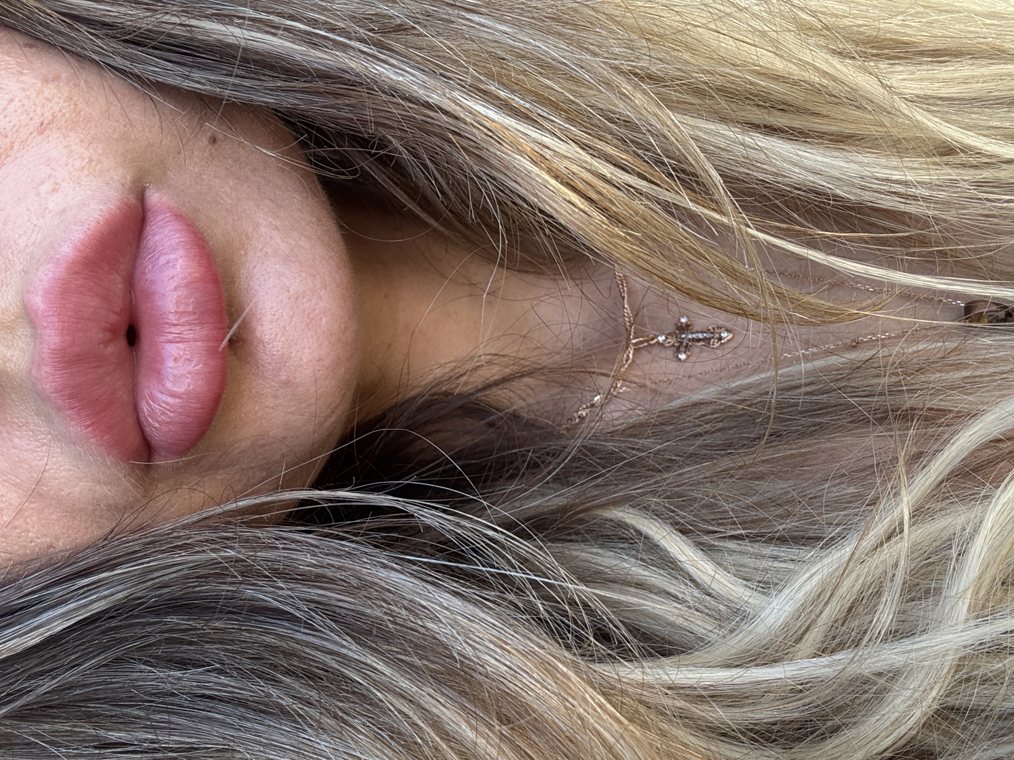 A close-up, high-angle shot captures a portion of a person's face, primarily focusing on their mouth and a cascade of hair. The subject's lips are full and pink, slightly parted with a small dark opening visible between them. A tiny white, hair-like strand rests near the lower lip. Above the upper lip, a small, dark mole is visible on the skin, which appears smooth.
The person's long, wavy hair dominates much of the frame, flowing around the face. The hair displays a natural mix of colors, ranging from lighter blonde or light brown tones to darker, possibly graying or shadowed strands, creating a textured appearance. Partially visible amongst the hair on the right side of the frame is a delicate, gold-toned necklace. It features a small, ornate cross pendant embellished with tiny, sparkling stones, likely diamonds or crystals. The lighting is bright, suggesting an outdoor or well-lit indoor setting during the daytime. The perspective indicates the person may be lying down or the photo is taken from above, with the hair spread out. There is no visible text in the image. The image was captured in Saint Petersburg, Russia.