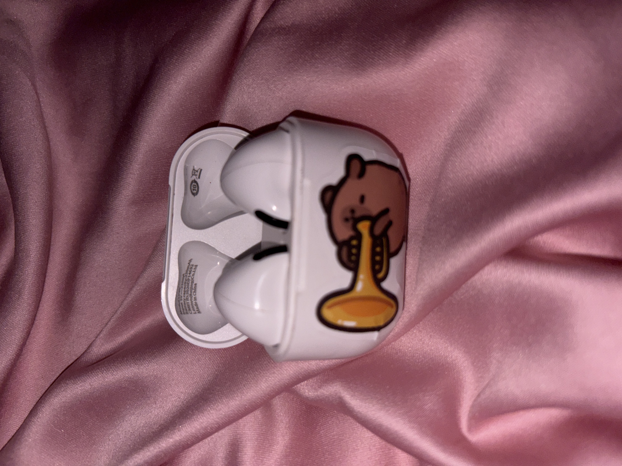 An open white wireless earbud charging case with a cartoon bear playing a trumpet on the front is placed on a pink satin fabric. The case is positioned in the center of the frame, with the lid open to reveal two white earbuds inside. The fabric has visible folds and creases, creating a soft, draped effect. The lighting is dim, casting shadows on the fabric and highlighting the glossy texture. No city or location information is available.