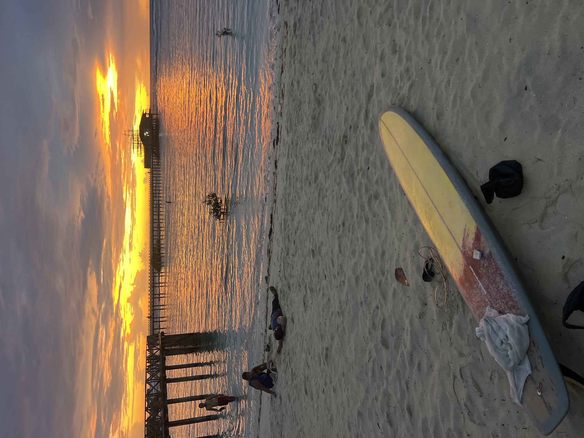 In General Luna, Philippines, a serene beach scene unfolds at sunset, dominated by the warm hues of the setting sun reflecting on the ocean. The sky is a canvas of vibrant oranges and yellows, with soft clouds tinged by the twilight. A wooden pier extends into the water, leading to a unique, multi-story structure built on stilts, resembling a rustic boat with scaffolding. The water is calm, with gentle ripples catching the sunset's glow.
On the sandy shore, several individuals are present. In the shallow water near the pier, a young person stands, appearing to be wading. Further down the beach, two individuals are seated on the sand. One is leaning back, resting on their arms, while the other is sitting upright, looking towards the water. Another person is lying down on the sand, appearing to be asleep or resting. In the foreground, a yellow surfboard with red and white accents lies on the sand, a towel draped over its tail. Beside the surfboard are a black bag and what appears to be a leash or cord. The sand is smooth, with visible footprints and scattered leaves. Small mangrove-like plants are visible emerging from the water near the shore.