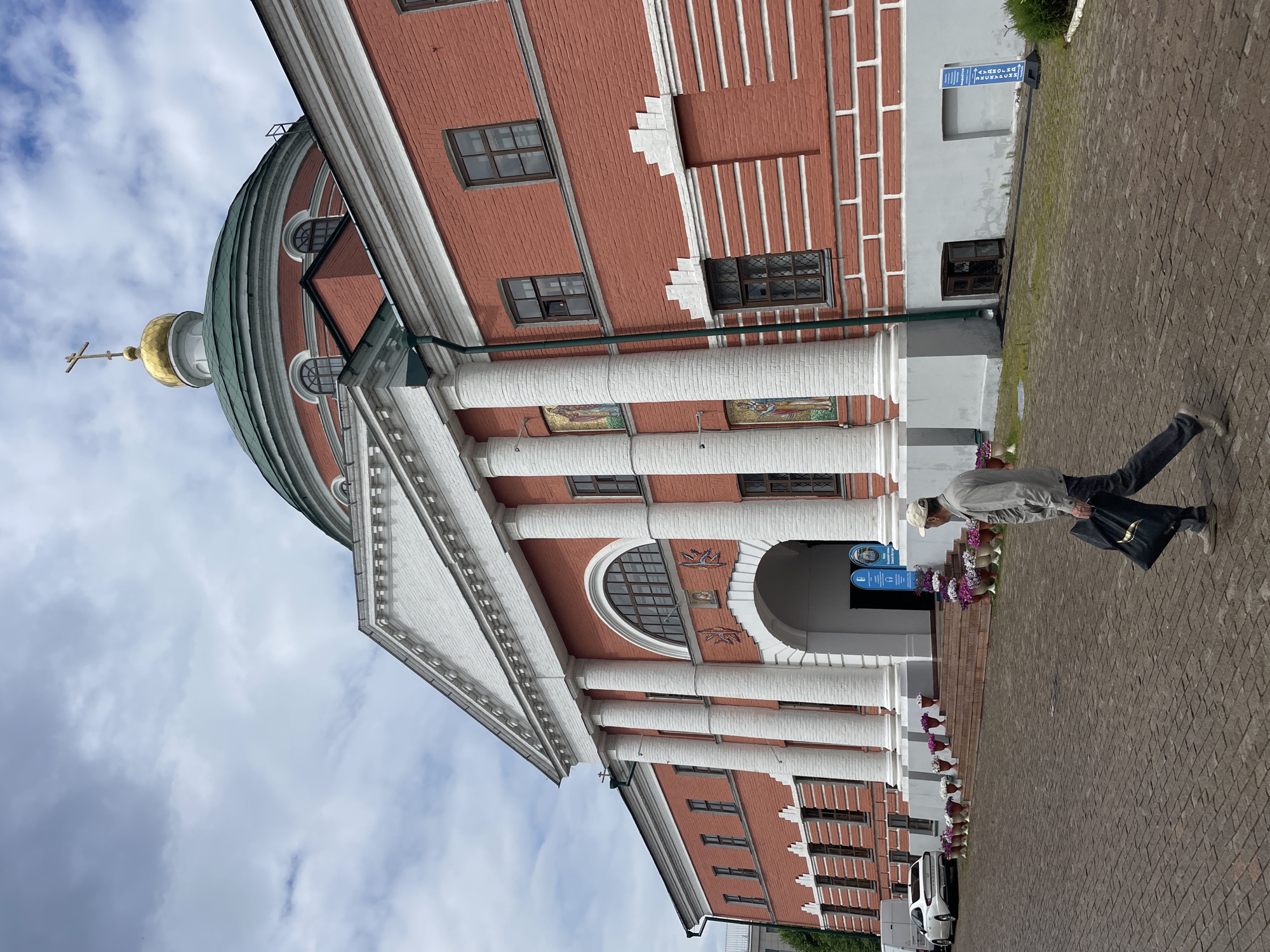 The image shows the exterior of the Cathedral of Saints Peter and Paul in Kazan, Russia. The cathedral is a prominent red brick building with white columns and a green dome topped with a golden cross. A man in a light-colored shirt and a baseball cap is walking across the cobblestone courtyard in the foreground, carrying a black bag. He is walking from right to left. The sky is overcast with scattered clouds. Several cars and planters with flowers are visible near the entrance of the cathedral. The overall scene depicts a cultural and religious landmark on a typical day.