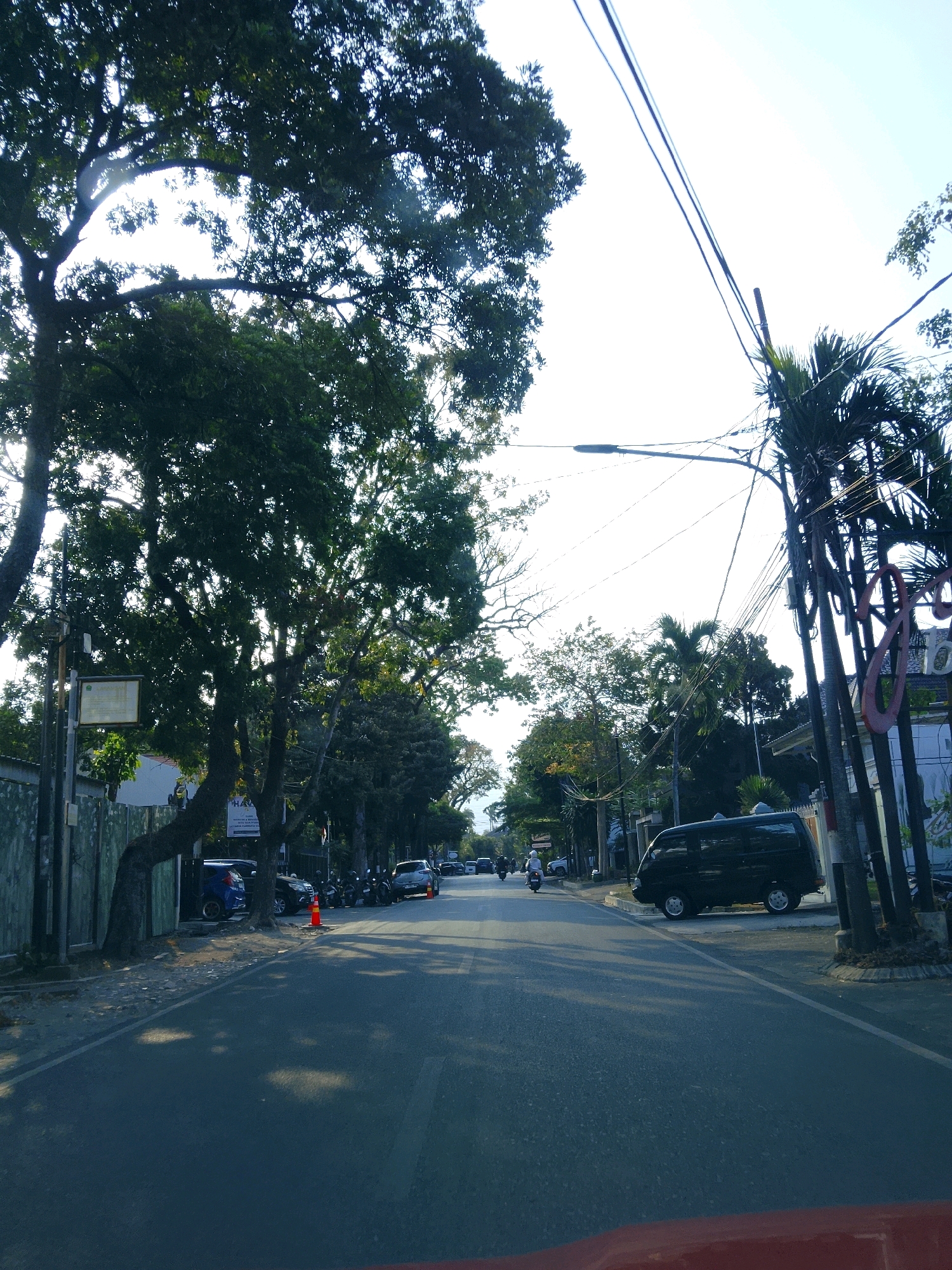 The image depicts a street scene in Malang, Indonesia, likely during daylight hours with the sun partially obscured by trees. The road is asphalt, with a white dividing line visible. Several vehicles are parked along the side of the road, including a dark-colored minivan. Some motorcycles and a scooter are also present, with one person on a scooter visible in the distance.
On the left side of the road, a tall tree with dense foliage casts shadows on the pavement. A fence runs along the side, bordering what appears to be a property. A street sign or information board is mounted on a pole near the tree.
On the right side of the road, a sign with large, stylized red letters is visible, along with palm trees and power lines. The sky is bright and appears clear, with some evidence of atmospheric haze. The overall impression is of a residential or commercial street with moderate activity. The presence of specific architecture and vegetation, combined with the context of Malang, Indonesia, helps to pinpoint the location.