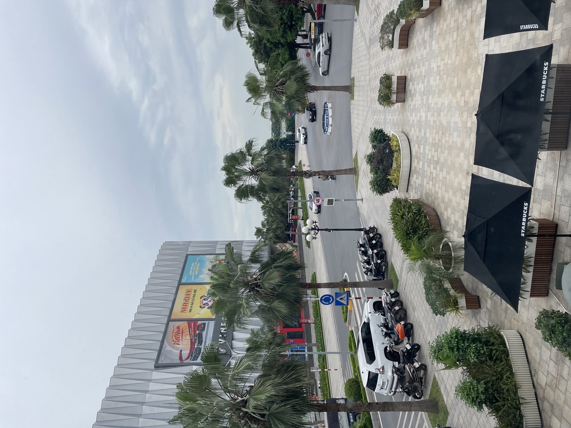 The image depicts a modern urban scene in Hanoi, Vietnam, likely within a commercial or entertainment district. In the foreground, several black Starbucks umbrellas are partially visible, set up on a paved area with planters containing greenery. Further back, a white SUV is parked alongside a row of motorcycles. Beyond the vehicles, a multi-lane road curves through the scene, with several cars and motorcycles in motion.
A large, modern building dominates the left side of the image, featuring a distinctive geometric facade and large billboards advertising "Vietnam," "MR.DIY," and "XANH GÌ LẠI." Palm trees are scattered throughout the scene, adding a tropical element to the urban landscape. The sky is overcast, suggesting a cloudy day. The overall impression is one of a bustling, contemporary city environment with a blend of commercial activity, transportation, and green spaces.