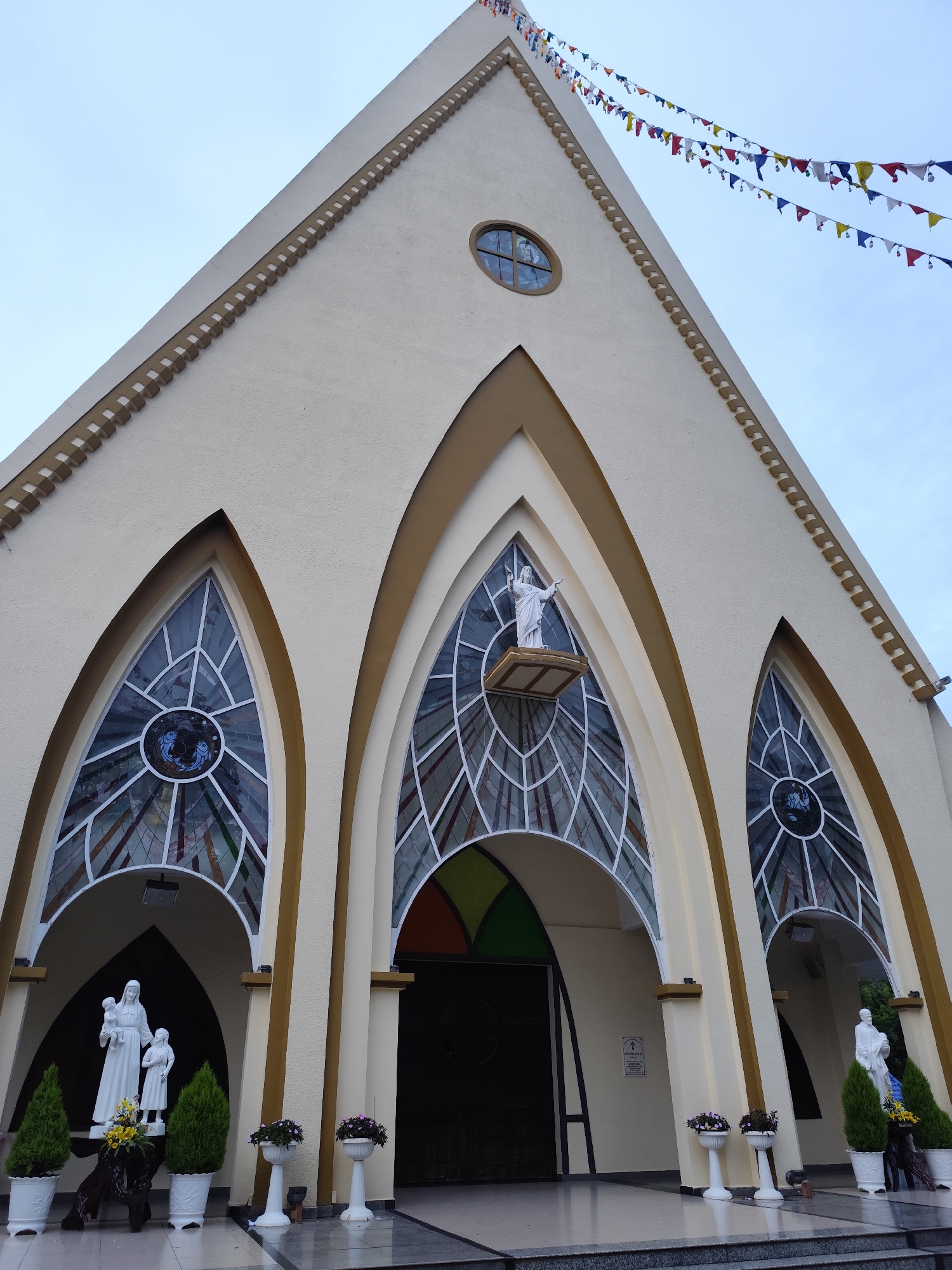 The image shows the facade of the Domaine de Marie church in Da Lat, Vietnam. The church features a cream-colored exterior with three prominent arched entrances. Stained-glass windows adorn the arches, with the central arch containing a statue of a saint positioned above the doorway. Statues of Mary and other saints are located on either side of the entrances. Small trees and flower pots line the entrance area. A string of colorful triangular flags decorates the roofline.