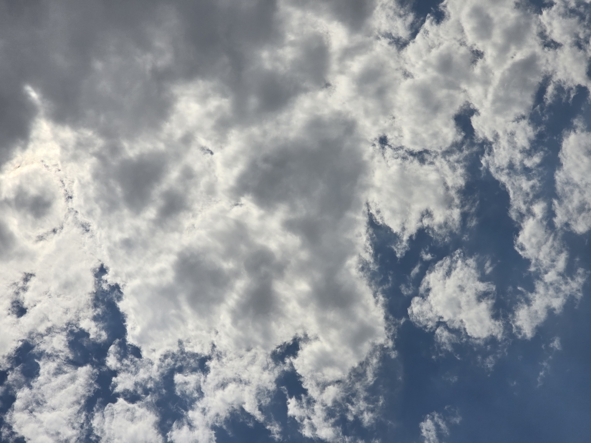 A vertical, medium shot captures a sky filled with a mix of white and gray clouds against a backdrop of blue sky. The clouds are varied in texture and density, with some appearing fluffy and bright, while others are darker and more imposing. Sunlight is visible filtering through the clouds in the upper right portion of the image, creating bright, illuminated patches and a hazy effect. The lower portion of the image shows more distinct blue sky interspersed with white clouds. There are no people, subjects, objects, or actions depicted. The scene is an outdoor, natural environment, specifically the sky above Podgorica, Montenegro.