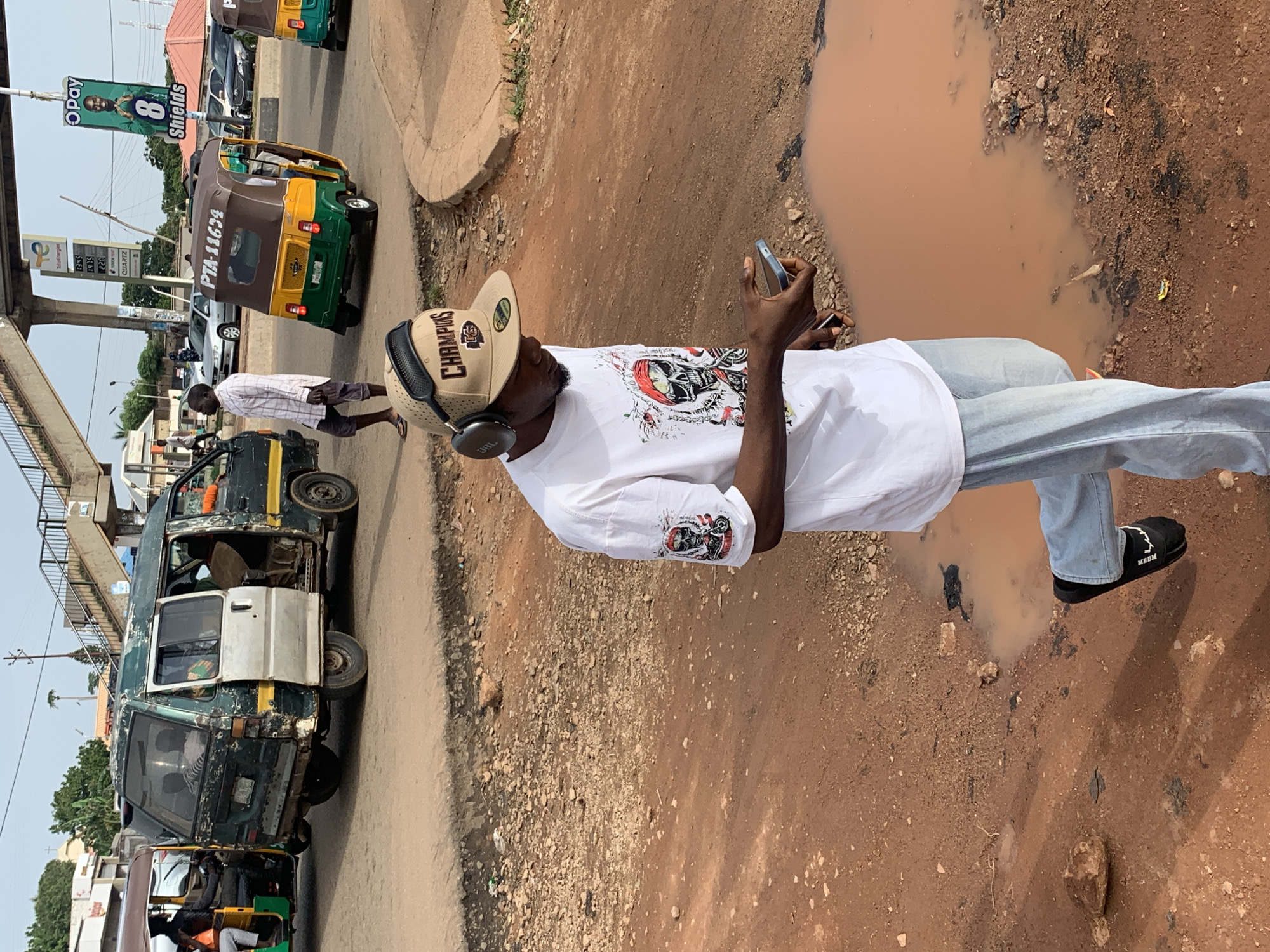 The image depicts a street scene in Gura Top, Nigeria, with a focus on a young man walking while looking at his phone and wearing headphones. He is dressed in a white t-shirt with a graphic print on the sleeve, light blue jeans, and black open-toed shoes. He is also wearing a tan baseball cap with "CHAMPIONS" printed on it. Behind him, a dilapidated green and silver van is parked, its doors open, revealing passengers inside. To the right of the van, a yellow and green tricycle, a common mode of transport in Nigeria, is visible. The ground is a mix of reddish-brown dirt and gravel, with a large muddy puddle in the foreground. In the background, a pedestrian bridge and various buildings are present, suggesting an urban environment. Several other vehicles, including cars and another tricycle, are also on the road. The lighting suggests it is daytime.