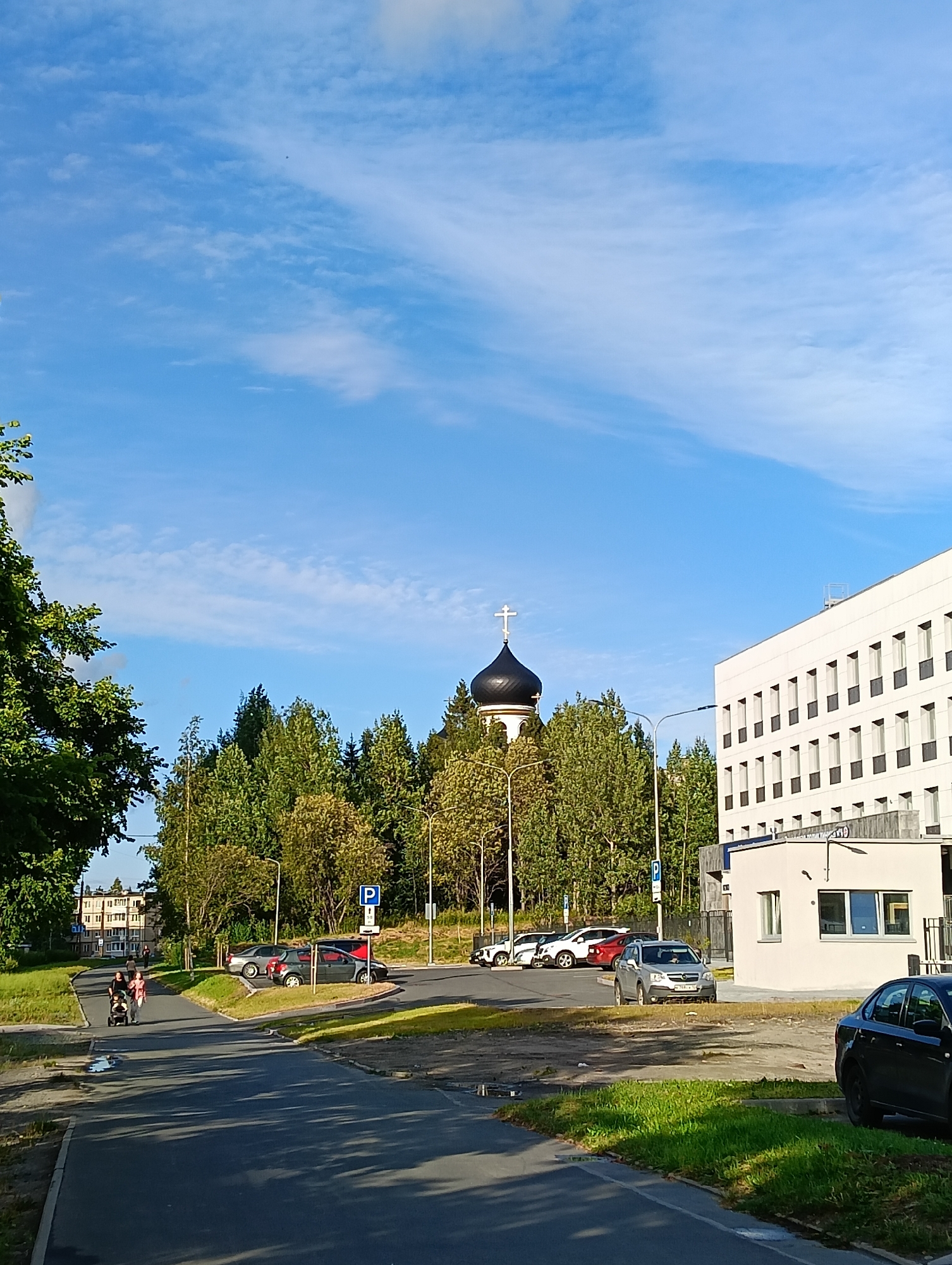 A street view captures a bright, sunny day in Petrozavodsk, Russia. In the foreground, a paved road leads towards a parking lot filled with several cars. The trees lining the street are lush and green. A white modern building with numerous windows dominates the right side of the frame. To the left, a dark-domed Orthodox church with a white cross on top stands out against the clear blue sky, surrounded by trees. A family with a stroller is seen walking on a sidewalk, further down the street. The overall atmosphere is serene, depicting a typical urban scene on a pleasant day.