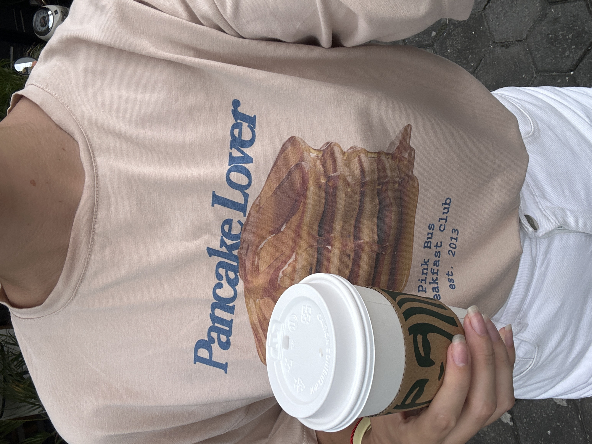 A person wears a peach-colored T-shirt with the words "Pancake Lover" and an image of pancakes printed on the front. The text "Pink Bus Breakfast Club est. 2013" is also visible. The person is holding a white coffee cup with a brown Starbucks sleeve. The person also wears white pants. The location is an urban area, based on the visible pavement. The city and region are unknown.