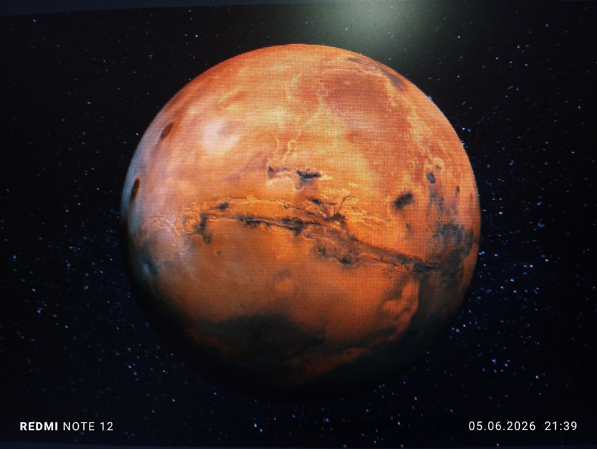 A photograph of a digital display screen depicts the planet Mars set against a dark, star-filled cosmic background. The prominent spherical object, rendered primarily in reddish-orange with darker, irregular geological features, displays a visible grid pattern across its entire surface and the surrounding space, characteristic of an LED or LCD screen display. The background is a deep blue-black field densely populated with numerous bright, pinpoint stars. Apparent lighting from the upper right illuminates the planet, creating subtle depth and relief on its surface. White text watermarks are present in the lower corners: "REDMI NOTE 12" is positioned in the bottom left, and "05.06.2026 21:39" is located in the bottom right. No individuals, specific actions, or other discernible objects are visible within the frame. This image is described as originating from the city of Burgas, Bulgaria.