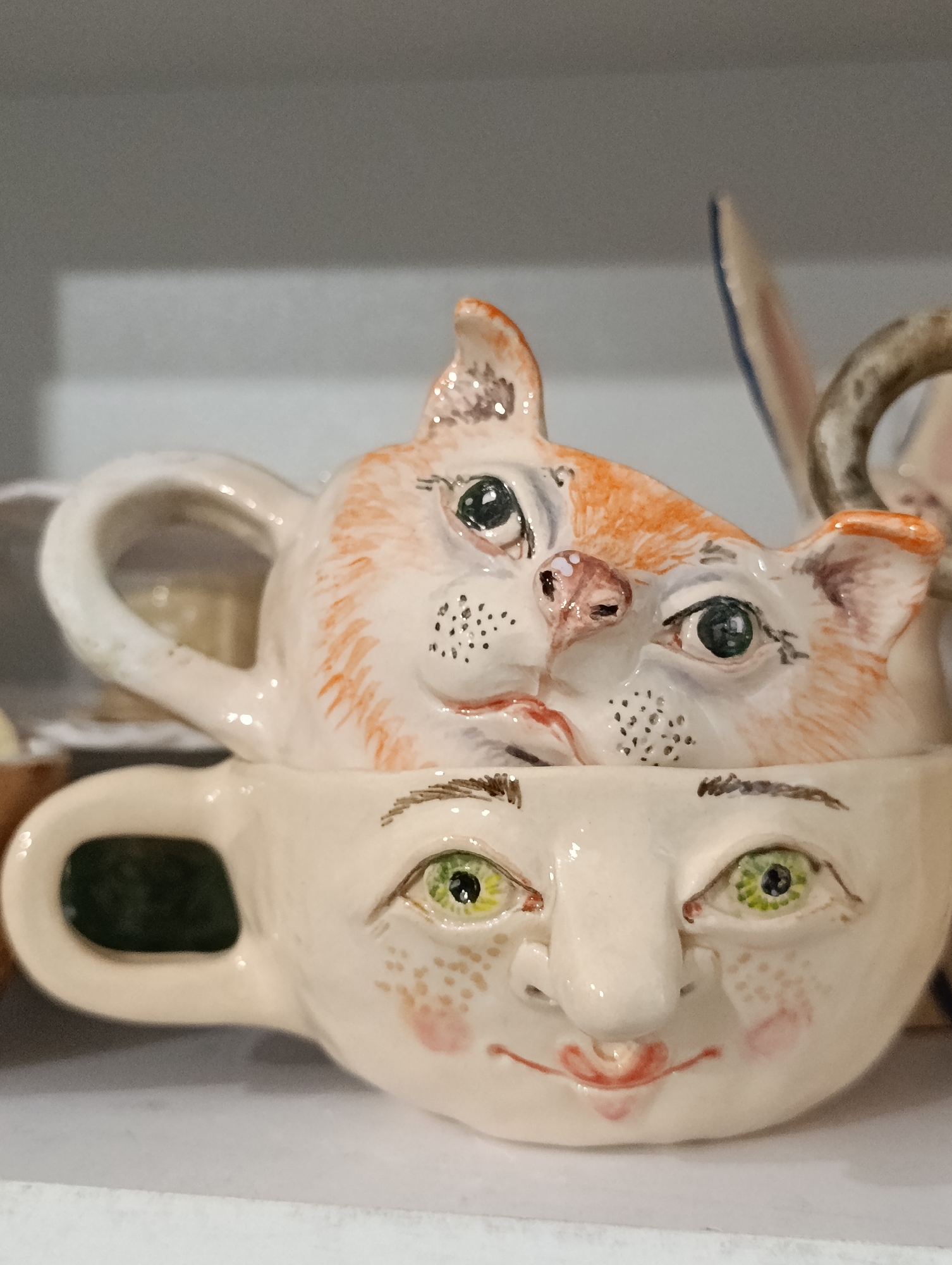 A ceramic cat's head, painted with orange markings and black dots on its cheeks, emerges from the top of a ceramic mug that has a human face painted on it. The cat's eyes are green and black, and its nose is pink. The human face on the mug has green eyes with black pupils, freckles scattered on its cheeks, and red lips. The mug has a handle on the left side, and the interior of the mug is dark green. The items are on a white shelf against a grey background. There is no identifiable information about the location in Bila Tserkva, Ukraine.