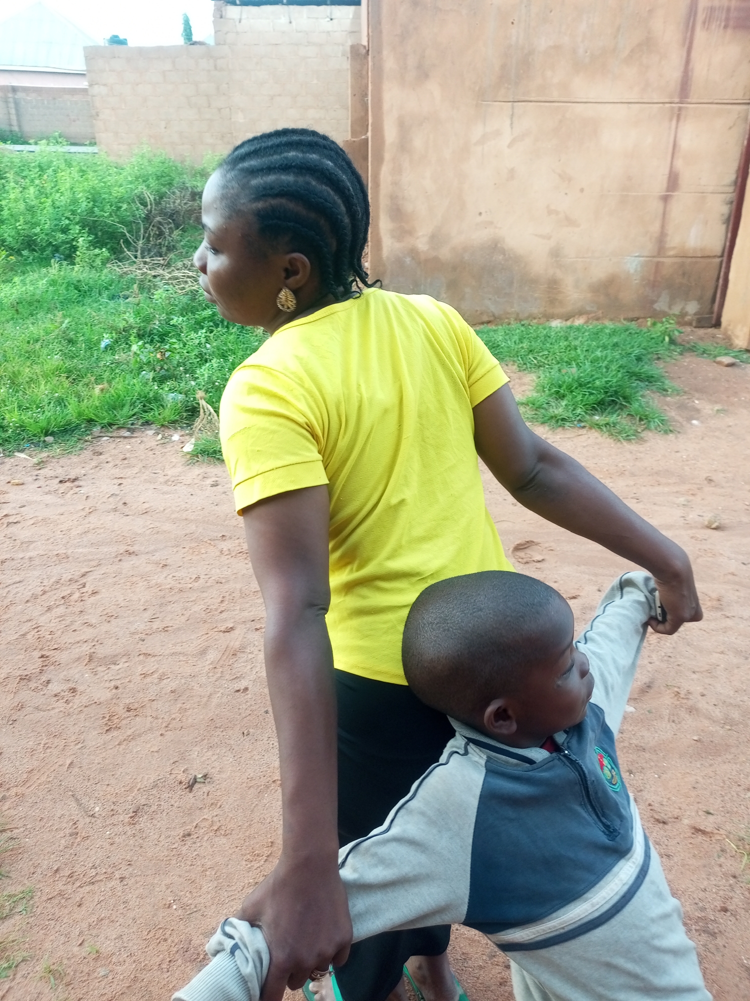 An adult female with braided hair and a yellow t-shirt stands outdoors in Tempure, Nigeria. She is holding the hands of a young child wearing a gray and blue jacket. The child is looking away from the camera. The ground is covered in sand and there is some grass and a building in the background. The scene appears to be a casual outdoor setting.