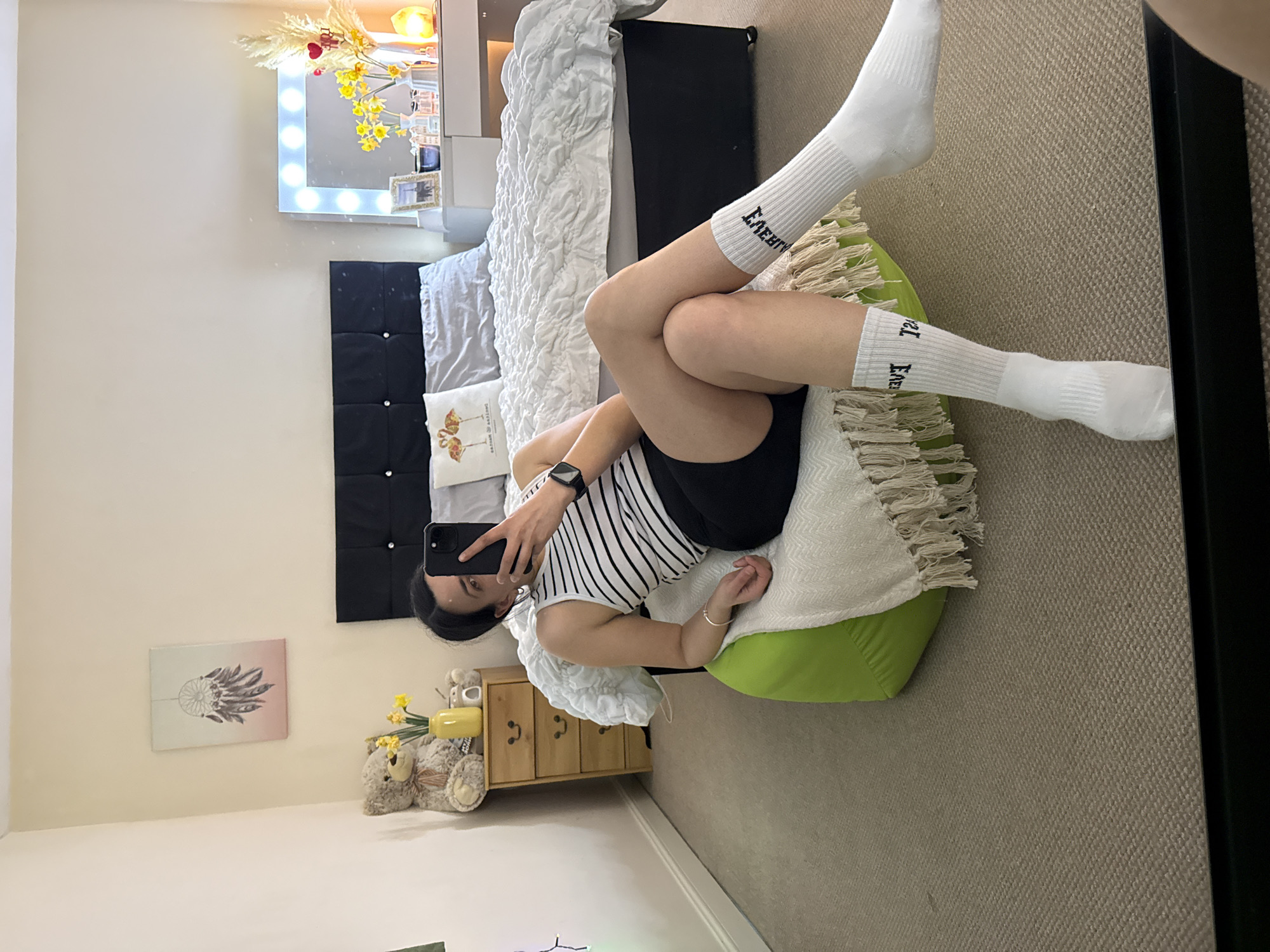 The image shows a bedroom interior. A woman is visible, reclining on a lime green beanbag chair, wearing white socks with black lettering, black shorts, and a black-and-white striped tank top. She is holding a black smartphone in her left hand, presumably taking a selfie. The bedroom features a bed with a black headboard, a white comforter, and several pillows. A bedside table made of light wood sits next to the bed, adorned with teddy bears and a yellow vase containing flowers. On the wall, there is a picture of a dreamcatcher and a vanity mirror with lights. The location is not inferable from the image.
