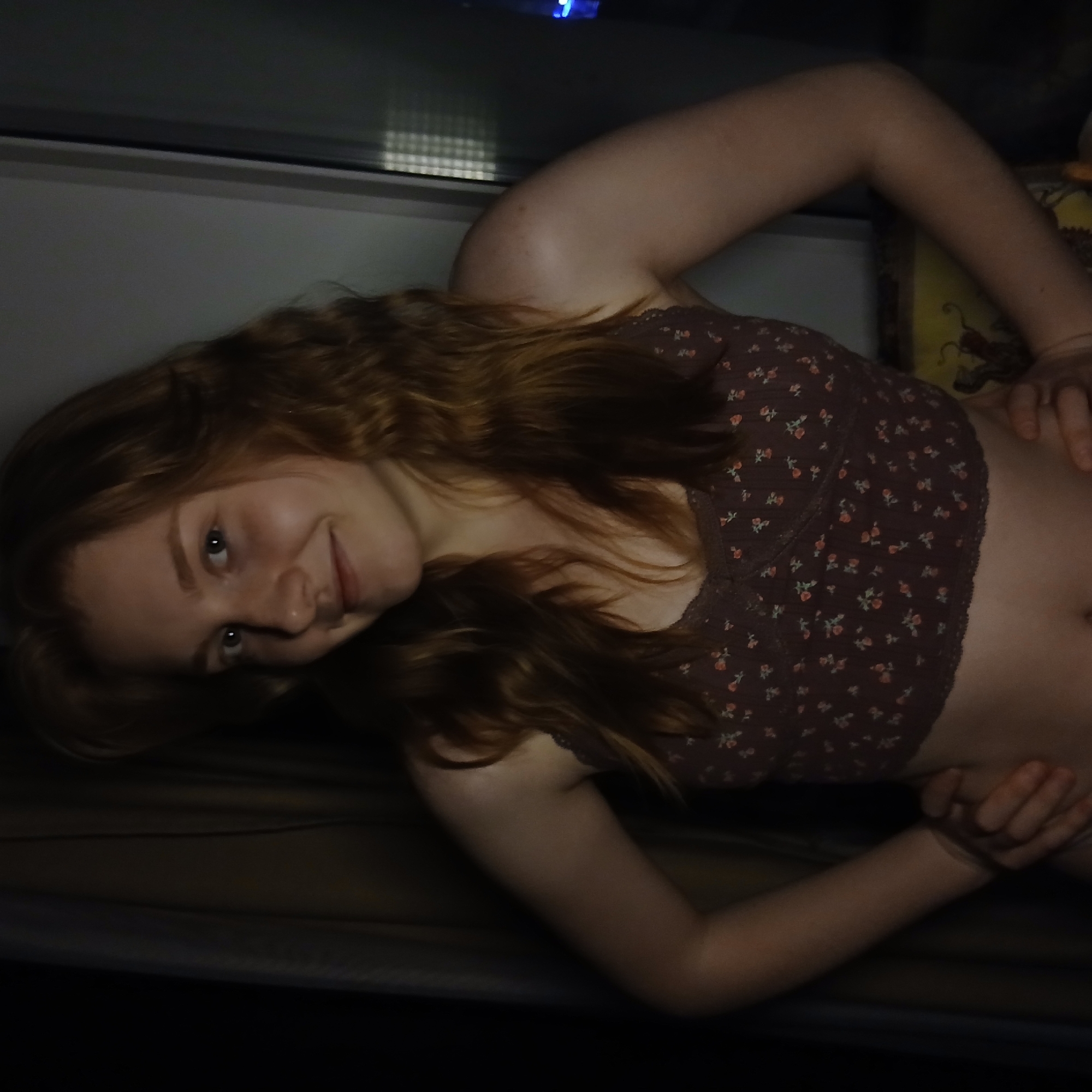 A young woman with long, reddish-brown, wavy hair is depicted lying down at an angle, looking towards the viewer with a gentle, relaxed smile. She has fair skin and light-colored eyes. She is wearing a dark brown or maroon crop top adorned with small, lighter-colored floral patterns, revealing her midriff. Her arms are bent, with her hands resting near her hips.
The setting appears to be indoors, with dim lighting that suggests it is either evening or night. Behind her upper body, a window or reflective surface is visible, showing a subtle grid pattern, possibly from blinds or a screen, and a faint blue light in the upper background. The woman is resting on a dark-colored surface, likely a bed or sofa. A small portion of a yellow object with a darker pattern is visible behind her right arm. The overall atmosphere is calm and informal. Based on the provided context, the location is Yekaterinburg, Russia. There is no visible text in the image.