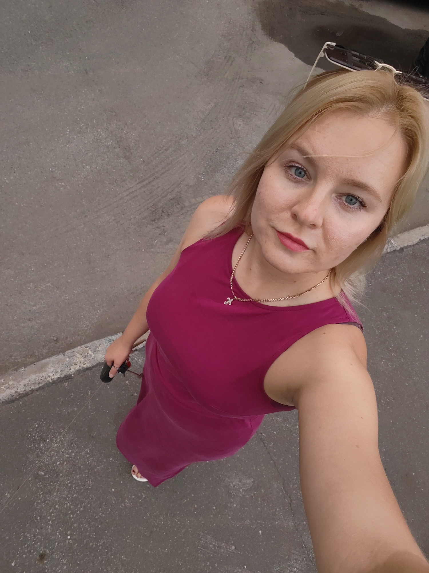 An overhead medium shot shows a blonde woman in a magenta dress standing outdoors. She is holding a black object in her right hand. Her left arm is visible. She has light blue eyes and pink lipstick. She is wearing a gold chain with a cross pendant. She is wearing white sandals. The background is a grey asphalt surface with a white concrete border. The location is Izhevsk, Russia.