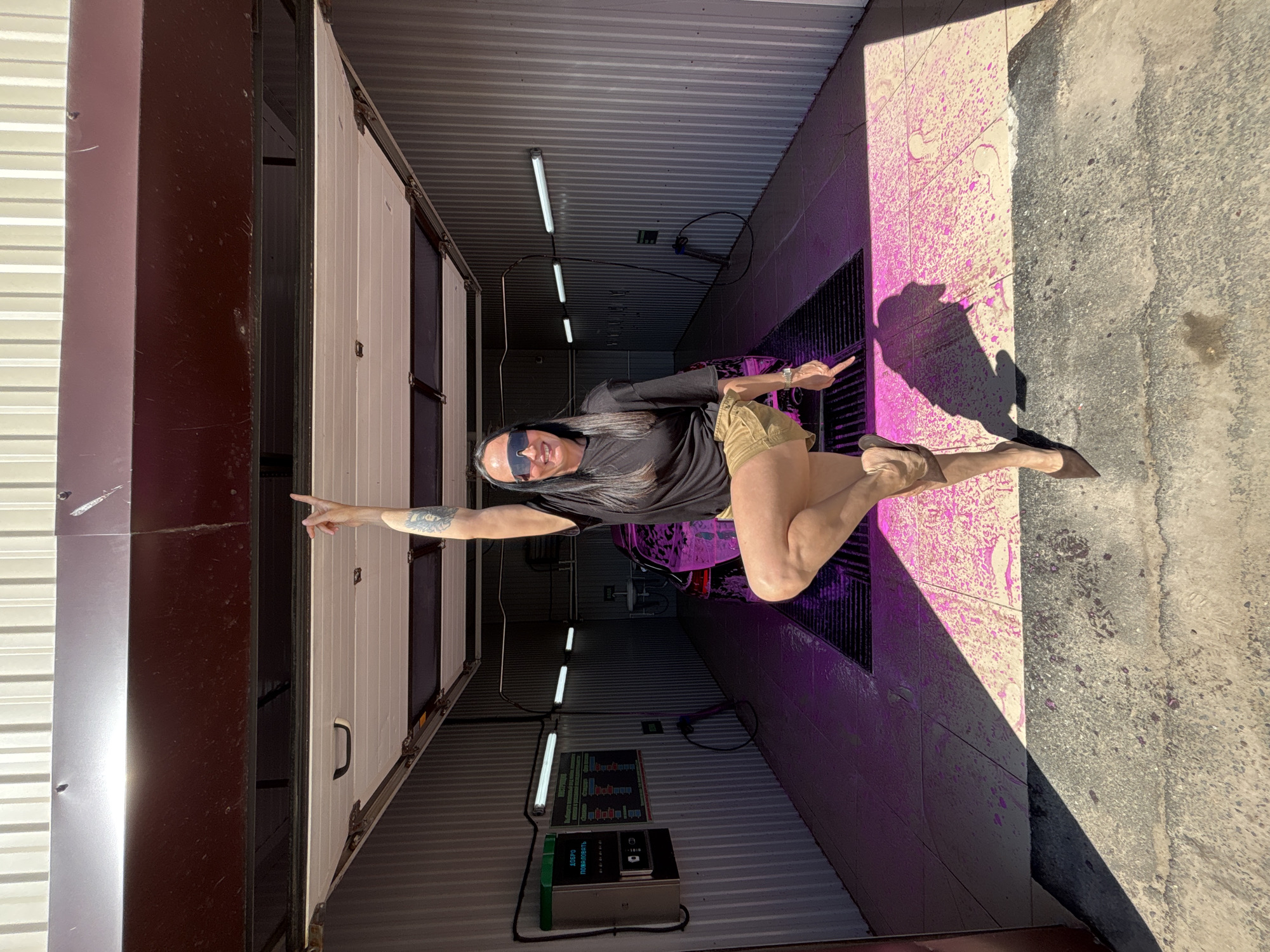 The image shows an individual standing in a car wash bay. The person has long dark hair, wears sunglasses, a black t-shirt, and khaki shorts. They are pointing with both hands, one toward the inside of the bay and the other toward the left side of the frame. The car wash bay has corrugated metal walls and ceiling. Fluorescent lights are mounted on the ceiling. A payment terminal is visible on the wall. The floor has a drainage grate. The image has a shadow cast across part of the scene, and pink streaks are visible on the walls, likely from car wash soap.
While specific location information isn't directly apparent in the image, it is stated that the location is in the city of Chaikovsky, Russia.