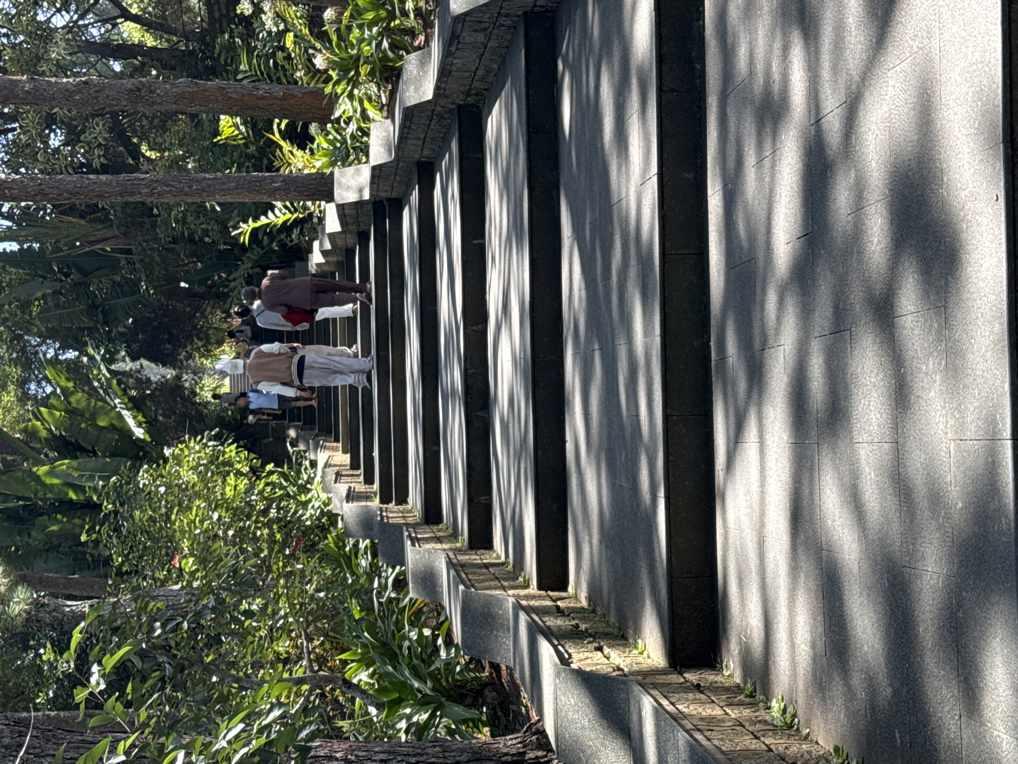 The image depicts a group of people ascending a wide, stone staircase in an outdoor setting characterized by lush greenery and tall trees. The staircase has multiple steps and is bordered by stone walls. Sunlight filters through the trees, casting dappled shadows on the steps. In the foreground, the staircase is empty, leading upwards into the dappled light. Further up, several individuals are visible, walking away from the camera and towards the top of the stairs. One person in the foreground is wearing a light-colored outfit and a head covering, followed by another individual in similar attire. Behind them, two more figures are walking up the stairs, one wearing a dark maroon outfit and the other in brown. This scene appears to be located at the entrance to the Thien Vien Truc Lam monastery, a prominent Zen Buddhist monastery in Da Lat, Vietnam, known for its picturesque setting and the long stone staircase leading up to its entrance.