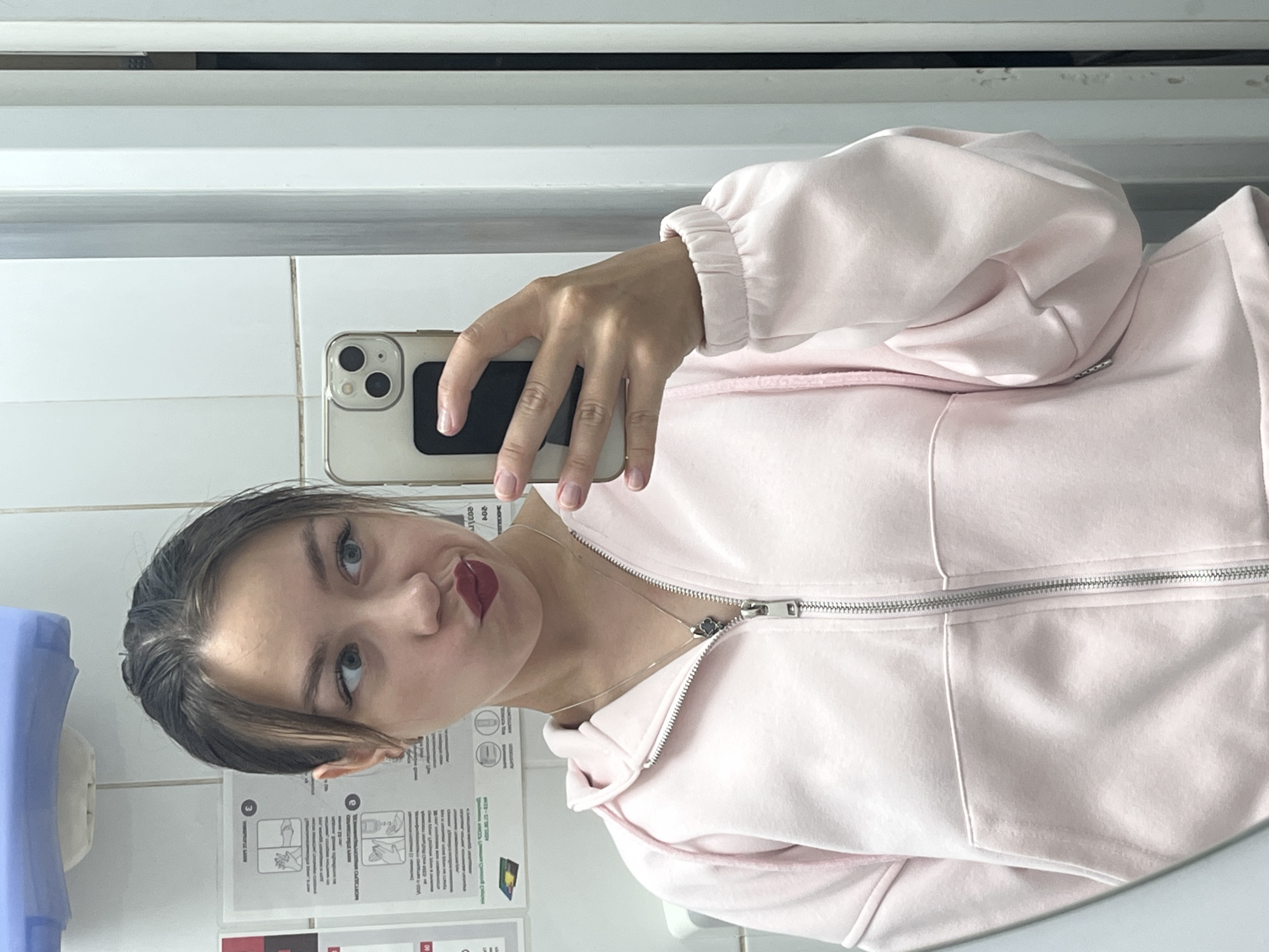 The image shows a young woman taking a selfie in what appears to be a public restroom. She is wearing a light pink zip-up hoodie and has dark hair pulled back. Her lips are pursed and she is wearing dark lipstick. She is holding a smartphone in her right hand, which is angled to capture her reflection in a mirror. Behind her, on the wall, are posters with instructional diagrams, possibly related to hygiene. The environment is tiled and appears to be a functional, rather than decorative, space. The specific location of Chaikovsky, Russia, is not determinable from the visual information presented in the image.