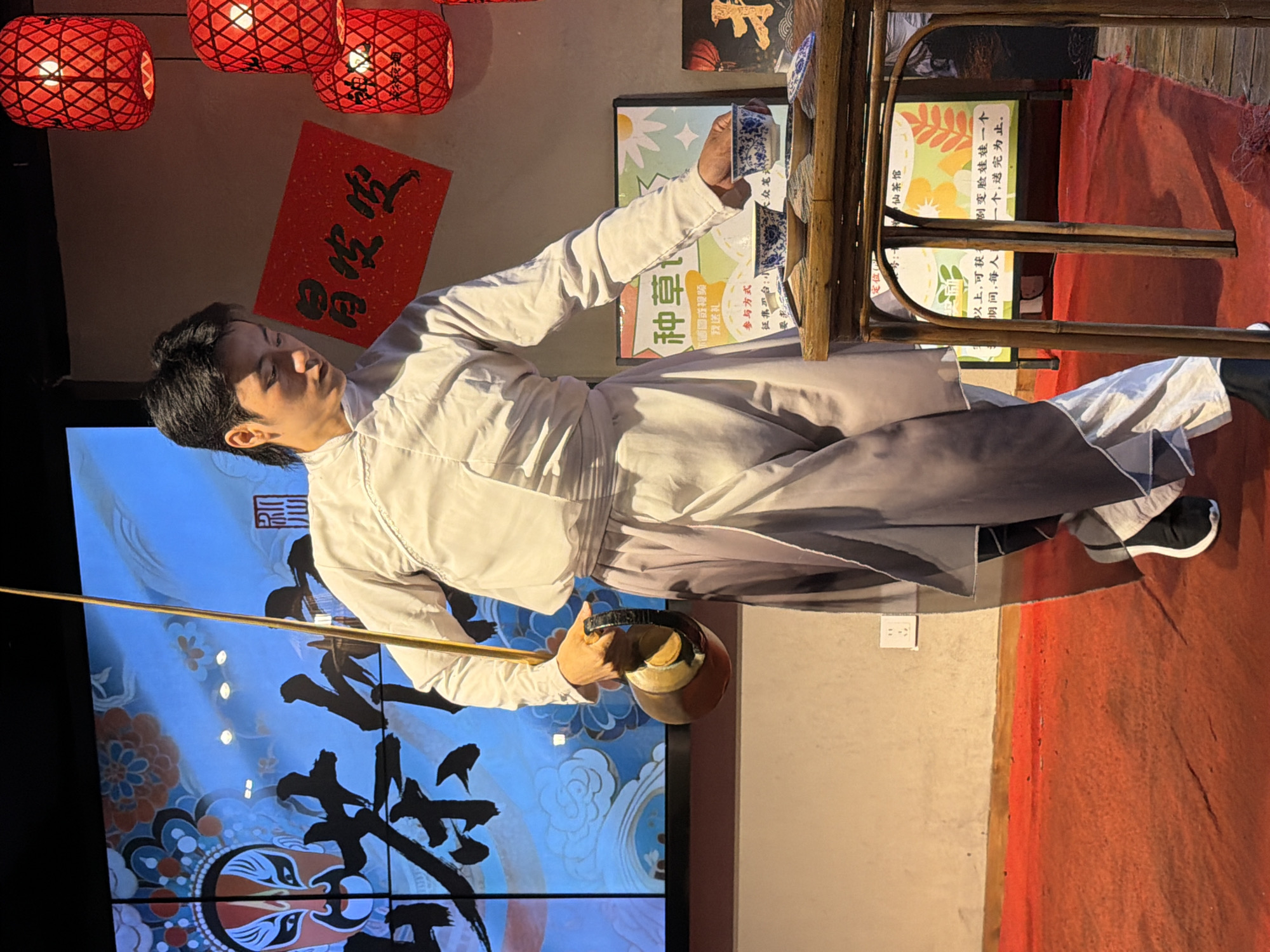 In Yuzhong District, China, a young man in traditional white and grey Chinese clothing is captured mid-performance. He holds a metal kettle with a black handle in his left hand and is reaching out with his right hand to pick up a blue and white patterned teacup from a wooden table. The table is set with several other similar teacups and saucers, and a small display board with Chinese text and floral imagery stands behind it.
The background features a large screen displaying a vibrant image of a Peking Opera mask and Chinese calligraphy, with red bamboo lanterns hanging from the ceiling on the right side of the frame. A red carpet covers the floor. The man's posture suggests a precise and graceful movement, typical of a tea ceremony or a theatrical performance.