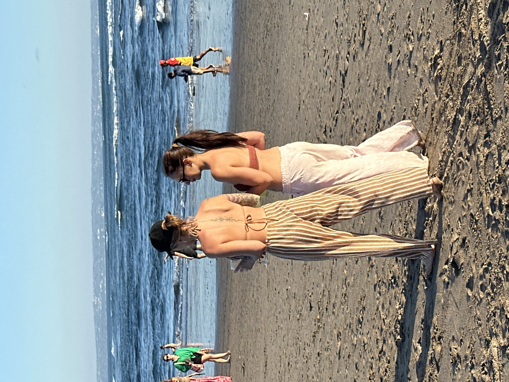 Two women stand on a sandy beach, facing away from the camera, looking out at the ocean. The woman on the left wears a black baseball cap, a string bikini top, and striped wide-leg pants. She holds a glass in her right hand. The woman on the right has her hair in a ponytail, wears sunglasses, a bikini top, and light-colored pants. In the background, other beachgoers are visible, including a person in a green shirt and shorts, and two figures, one in a yellow shirt and the other in blue, possibly near the water. The ocean has moderate waves under a clear blue sky. The scene is likely located on the coast of North Kuta, Indonesia, known for its beaches and surf breaks.