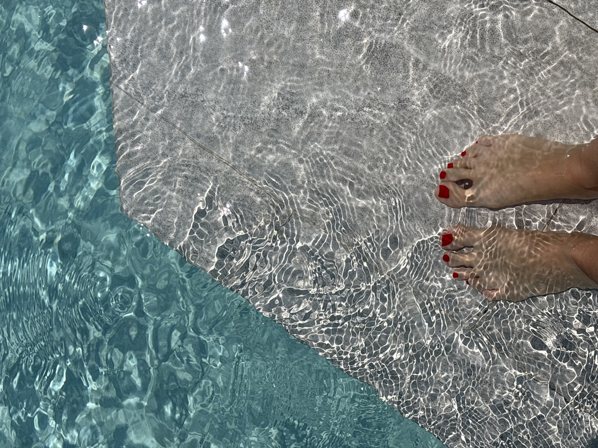 An overhead shot captures a person's feet partially submerged in clear, shallow water at the edge of what appears to be a swimming pool or a water feature. The feet, located in the upper right portion of the frame, are adorned with bright red nail polish on the toes.
The image is compositionally divided by a diagonal line where the water depth changes. On the upper right, a light-colored, textured surface, possibly pool tiling or concrete, lies beneath the shallow water. Intense sunlight refracts through the water, casting dynamic, shimmering patterns of light and shadow that ripple across this surface and the skin of the feet. The lower left section reveals deeper, turquoise-blue water, also displaying similar light patterns from the surface ripples.
The scene suggests a moment of relaxation or leisure on a bright, sunny day, indicated by the strong, direct sunlight. The clear water and serene setting evoke feelings of tranquility and cooling off. The overall aesthetic points to an outdoor, warm-weather environment, consistent with the Choeng Thale, Thailand location context. There is no visible text in the image.