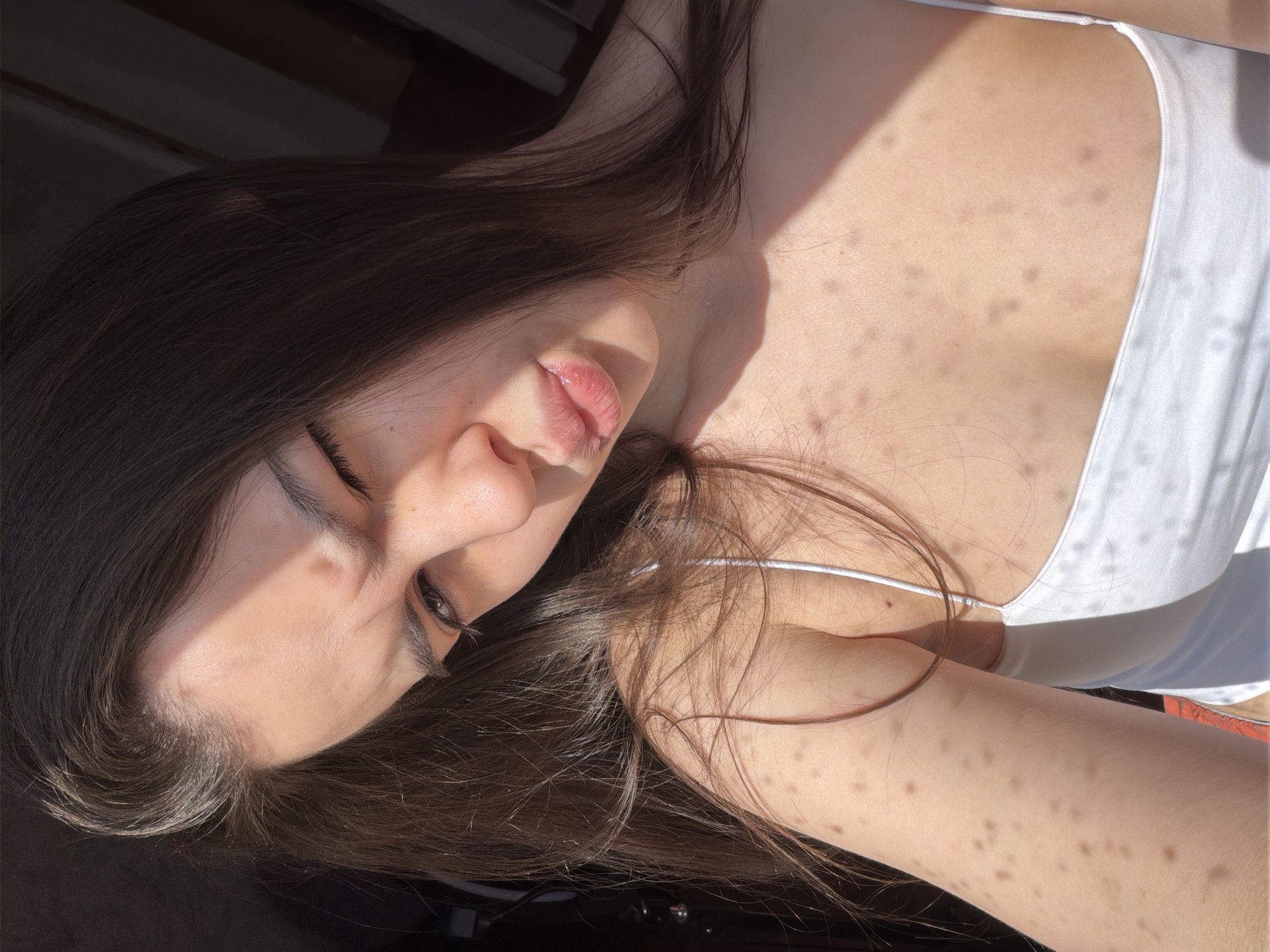 A close-up portrait shows a young woman reclining indoors, strongly illuminated by direct sunlight, casting defined shadows. Her head is tilted to the left, with her eyes mostly closed or looking downwards, and her lips slightly parted, conveying a serene expression. Long, dark brown hair frames her face and flows across her shoulders. She wears a simple white tank top with thin straps. Her skin, particularly on her chest, shoulders, and arm, is covered with numerous small, dark spots, which are emphasized by the bright, high-contrast lighting. The background is dark and out of focus, providing minimal environmental context beyond suggesting an indoor setting. There is no visible text. The location context provided for this image is Chaikovsky, Russia.