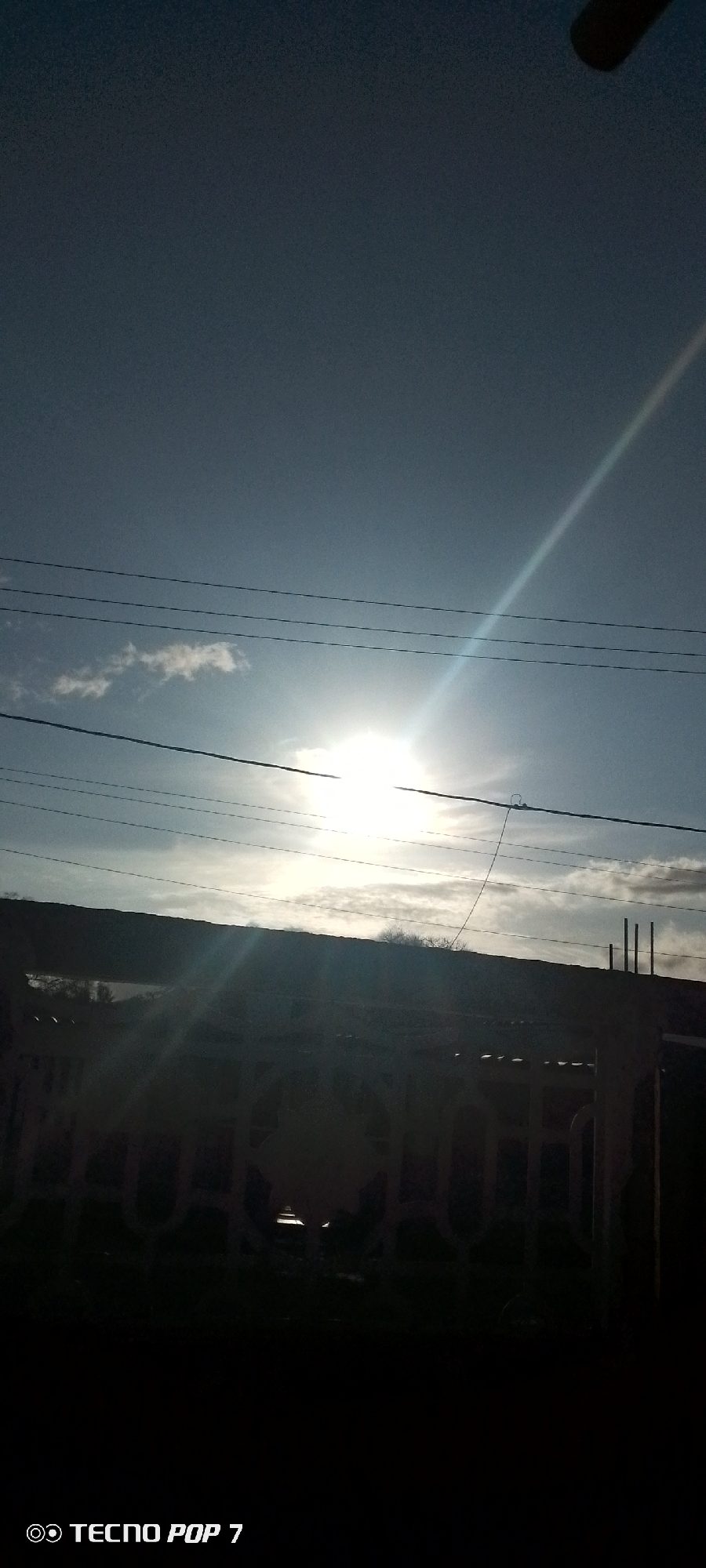 The image shows a view of Altagracia, Venezuela under a bright sky with scattered clouds. Power lines run across the frame near the center. Below the sky, there is a silhouette of a building or structure with what appears to be a fence in the foreground, partially obscuring the view. The sun is shining intensely, creating a lens flare effect. The lower portion of the image is in shadow. The camera model "TECNO POP 7" is visible at the bottom left corner.