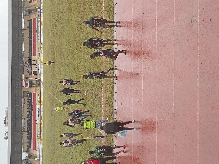 A sporting event is captured in an outdoor setting. A crowd fills stadium seating, visible on the left. On the field, individuals are scattered: some in athletic attire and others in security vests. To the right, a reddish-brown track stretches, marked with lanes, where several individuals are running or walking. The overall view is somewhat blurred, as if captured at a distance or in motion, with elements of a stadium and field clearly visible.