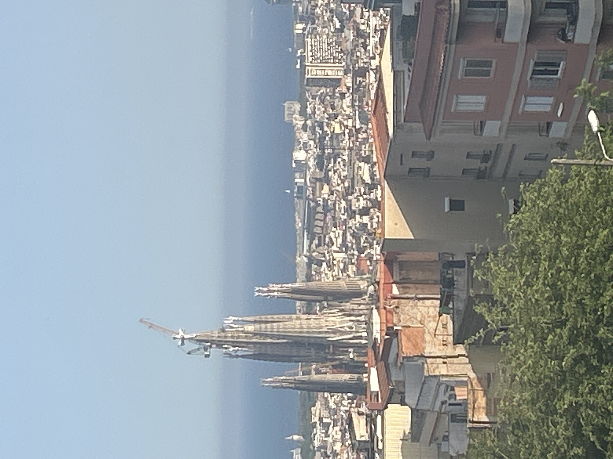 The image captures an elevated, panoramic view of a city, specifically Barcelona, Spain, extending towards the tranquil blue sea under a clear sky. Dominating the left side of the midground are the distinctive and intricate spires of the Sagrada Familia, with a construction crane visible next to one of them, indicating ongoing work. Surrounding this iconic landmark is a dense urban landscape comprised of numerous buildings, varying in architectural style, with some featuring red-tiled roofs and others presenting flat surfaces.
In the background, the sea stretches to the horizon. The foreground, particularly on the right, shows more city buildings and a large, leafy green tree partially obscuring parts of the view. A streetlight is also visible behind the tree. The scene is brightly illuminated by natural sunlight, suggesting a clear and sunny daytime. No individuals are visible, and there is no discernible text in the image. The composition conveys the grandeur of the city's architecture and its proximity to the Mediterranean coast.